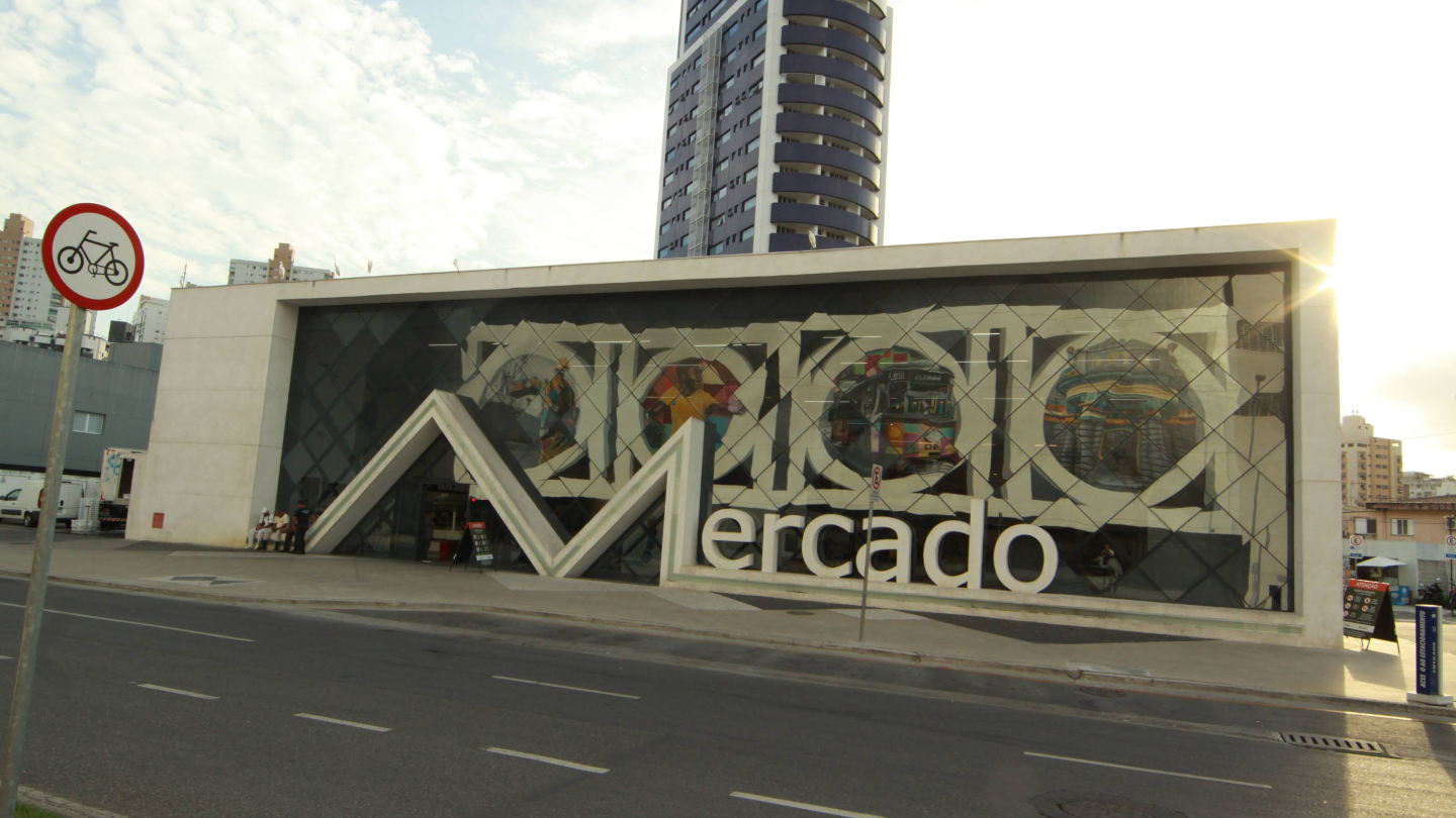 Fachada moderna do Mercado de Peixes de Santos, com grandes letras brancas que formam a palavra “Mercado” e painéis de vidro decorados com murais coloridos. À frente, passa uma avenida e, ao fundo, ergue-se um prédio alto e curvo.