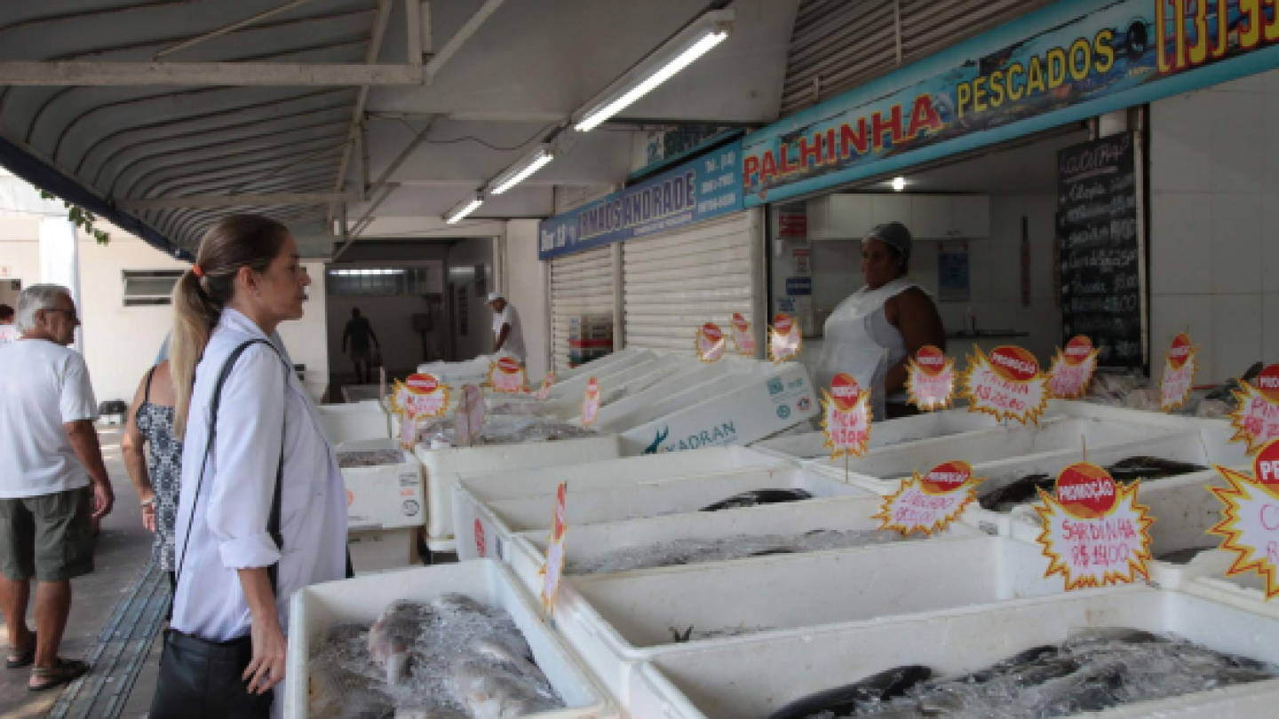 Mulher observa caixas de isopor cheias de peixes sobre gelo em um box do Mercado de Peixes de Santos, com placas de promoção destacadas
