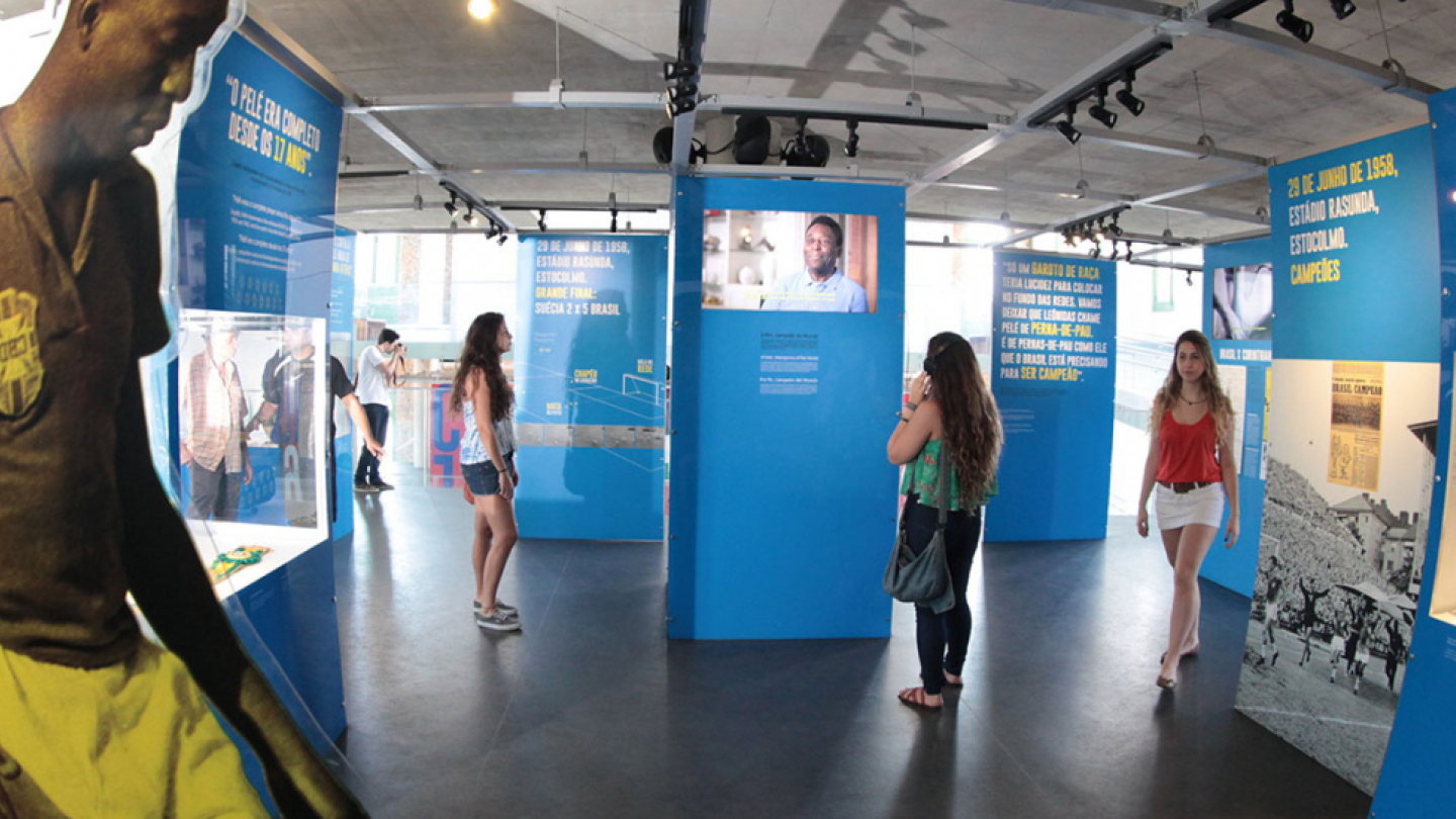 Interior de uma exposição sobre futebol: à esquerda, escultura em tamanho real de jogador com uniforme da Seleção Brasileira. No espaço, visitantes observam painéis azuis com textos, fotos e objetos históricos, em destaque a Copa de 1958.