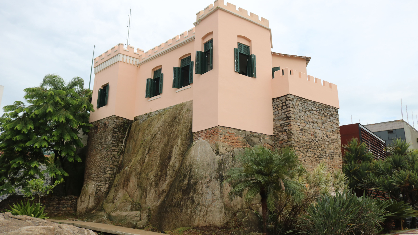 Um prédio histórico de cor salmão, com janelas de venezianas verdes e telhado com ameias. Ele está construído sobre um grande rochedo e cercado por vegetação