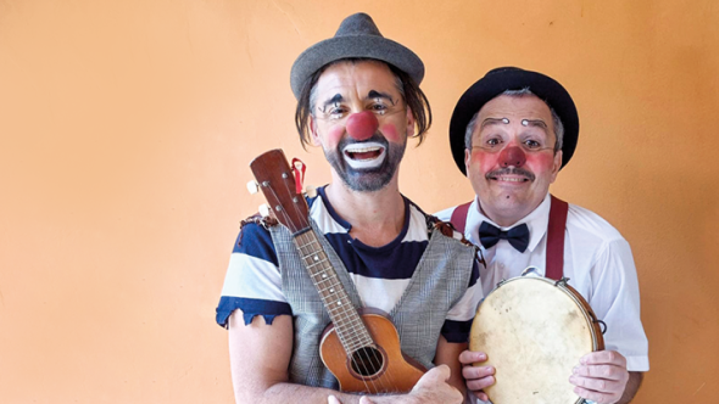 Dois palhaços sorridentes posam para a foto, um segurando um pandeiro e outro um cavaquinho
