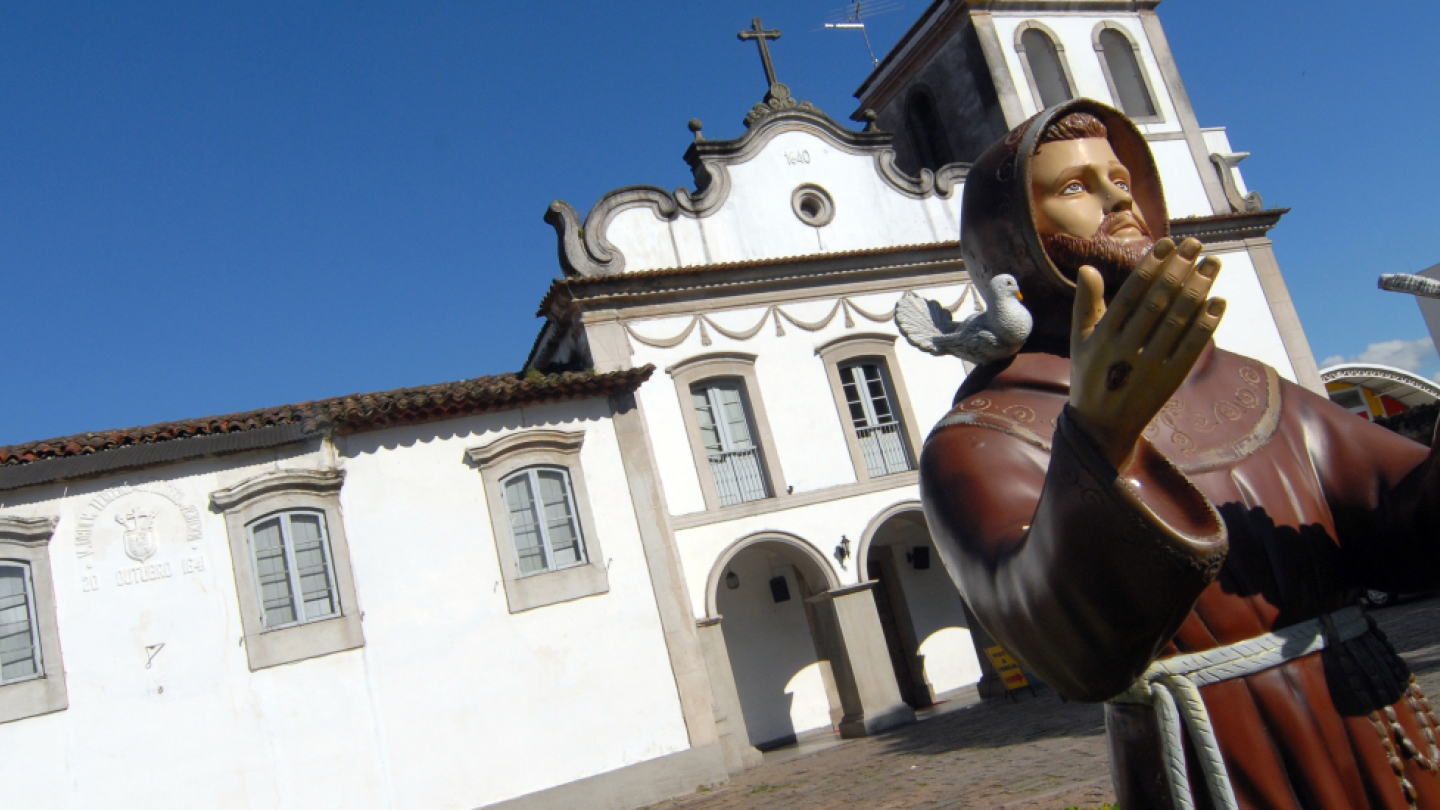A statue of Saint Francis of Assisi in the foreground, with white birds on his hands and shoulders. Behind him, under a clear blue sky, stands a white colonial-style church, with tiled roofs and a bell tower