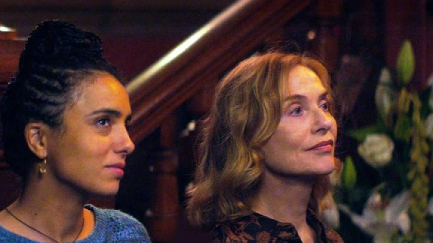 Duas mulheres em um ambiente interno. Elas estão posicionadas lado a lado, olhando atentamente para algo fora do quadro, à direita. A mulher à esquerda tem pele morena, usa os cabelos presos em tranças no alto da cabeça e veste uma blusa azul de tricô. Já a mulher à direita tem pele clara, cabelos loiros-acinzentados soltos, e veste uma blusa de tons escuros com estampa discreta.