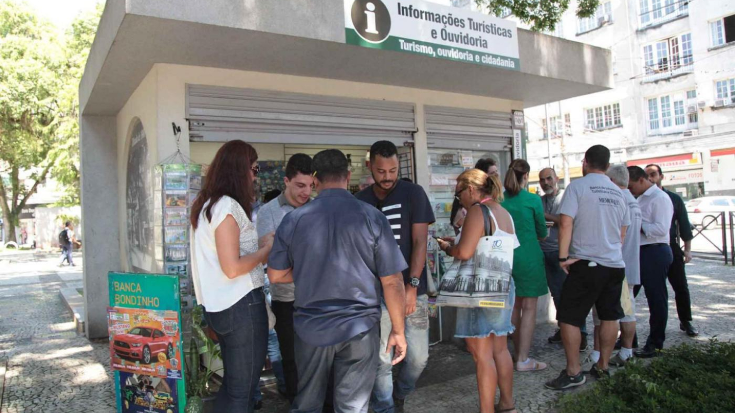 A imagem mostra uma banca de jornal localizada no Centro de Santos que também funciona como posto de informações turísticas e ouvidoria. Em frente ao espaço, um grupo de pessoas interage com os atendentes, demonstrando interesse pelos materiais disponíveis. O letreiro na parte superior indica claramente a função do local, voltada para turismo, ouvidoria e cidadania.