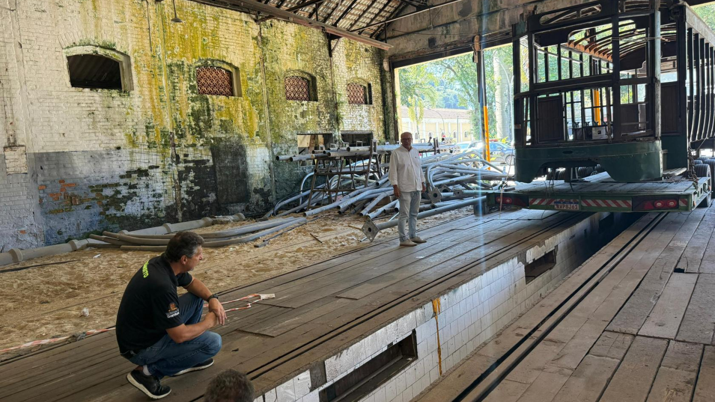 O ambiente é amplo, com piso de madeira e um fosso revestido de azulejos brancos, utilizado para manutenção dos bondes. Ao fundo, pela porta aberta, é possível ver árvores e parte da área externa, contrastando com o interior histórico do galpão.