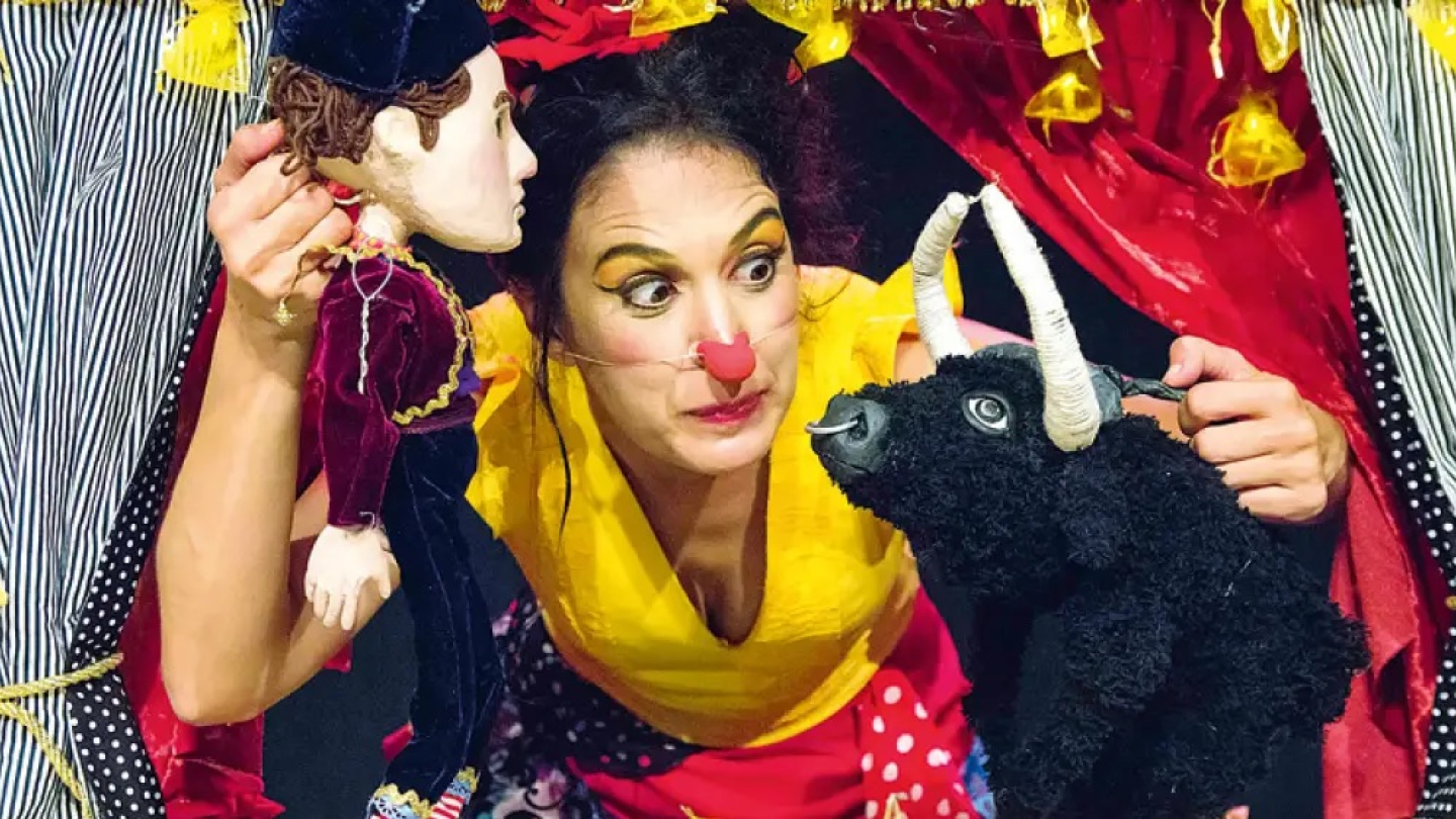 Atriz com nariz de palhaço interage de forma expressiva com dois bonecos, um toureiro e um touro, em cena colorida de teatro infantil