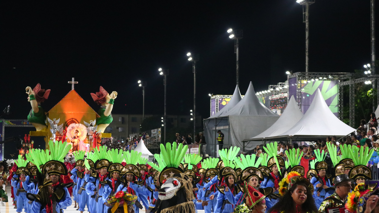 Desfile de carnaval à noite, com integrantes fantasiados em cores vibrantes, e um grande carro alegórico ao fundo