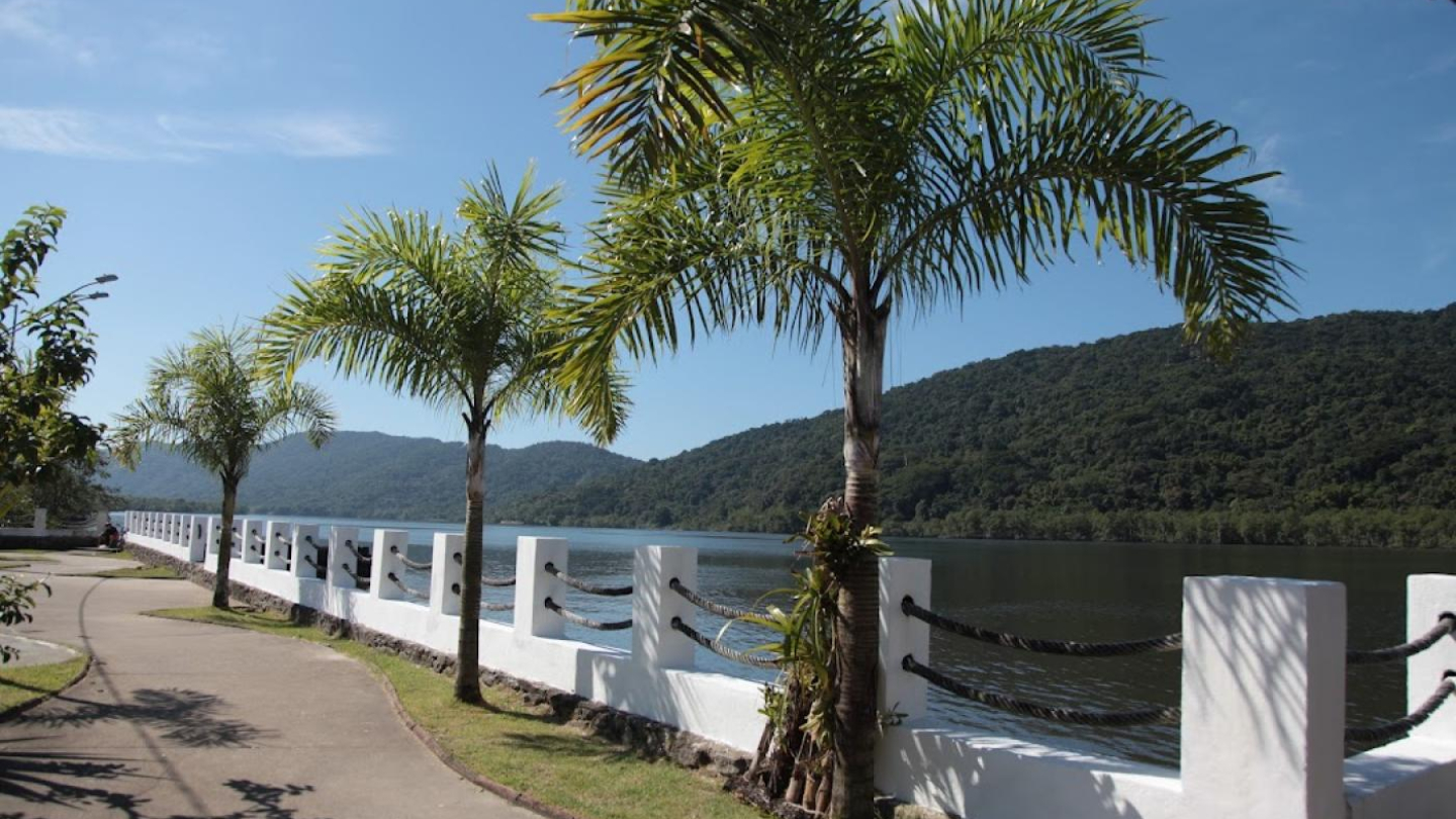 Calçadão arborizado com palmeiras, à beira do Canal de Bertioga, com vista para morros cobertos de vegetação da Mata Atlântica
