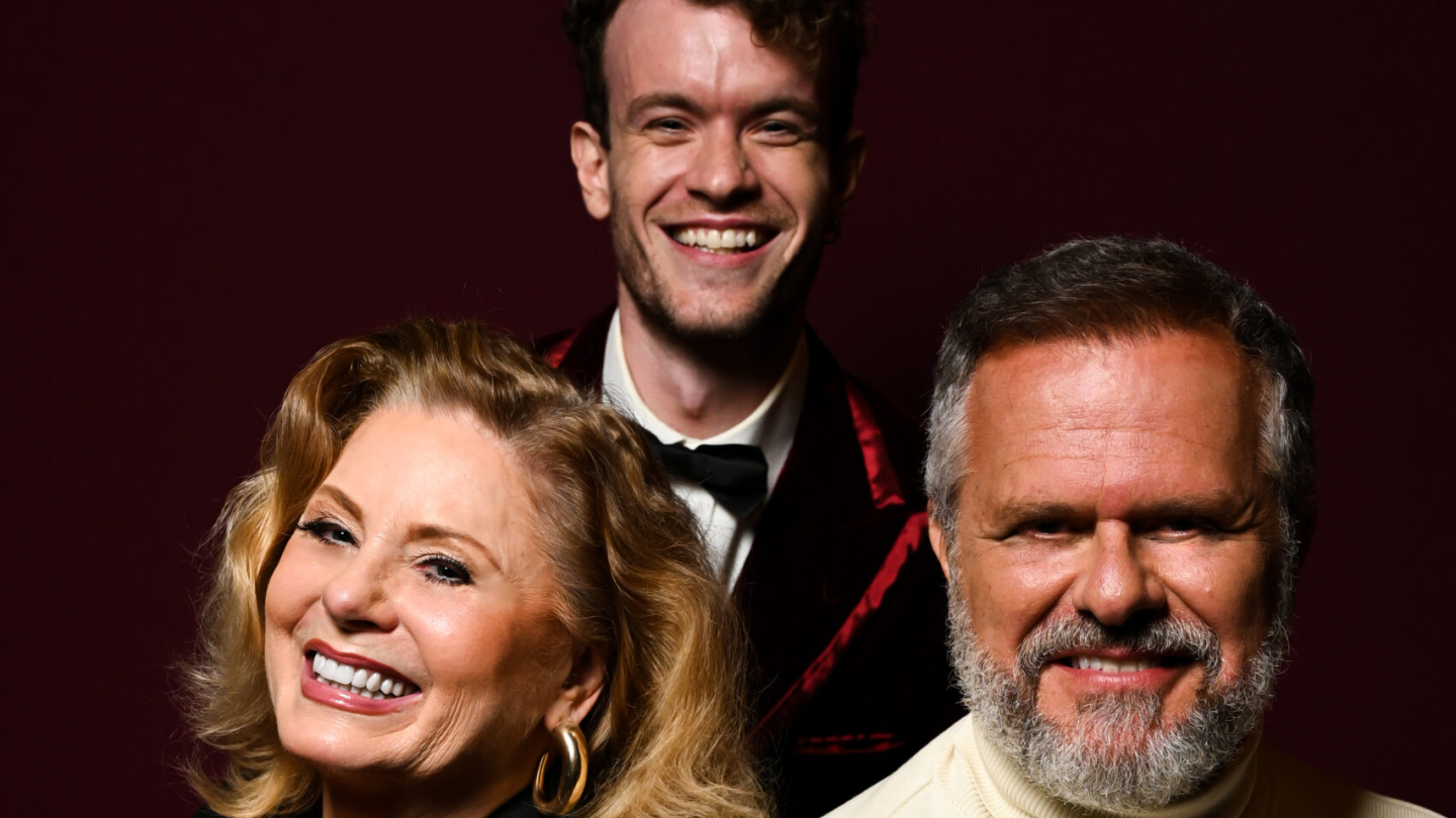 Uma mulher e um homem mais velhos estão em primeiro plano, sorrindo e olhando para a câmera. Um homem mais jovem, com cabelo cacheado e um laço borboleta, está em segundo plano, entre eles, também sorrindo