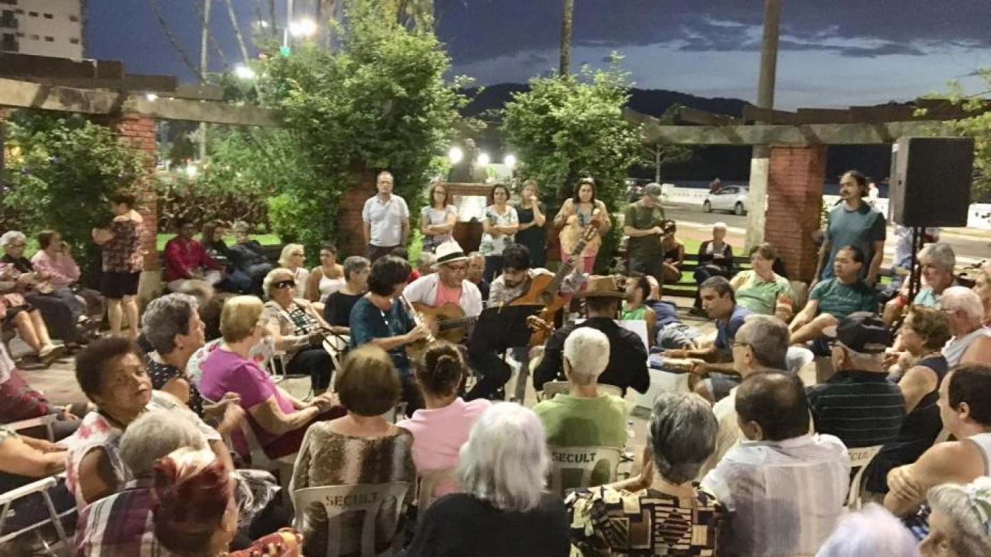 Grupo de pessoas reunidas ao ar livre, assistindo e tocando música em roda ao anoitecer