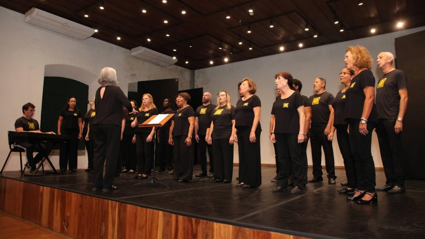 Coral com diversas pessoas em trajes pretos, perfilados no palco com iluminação discreta
