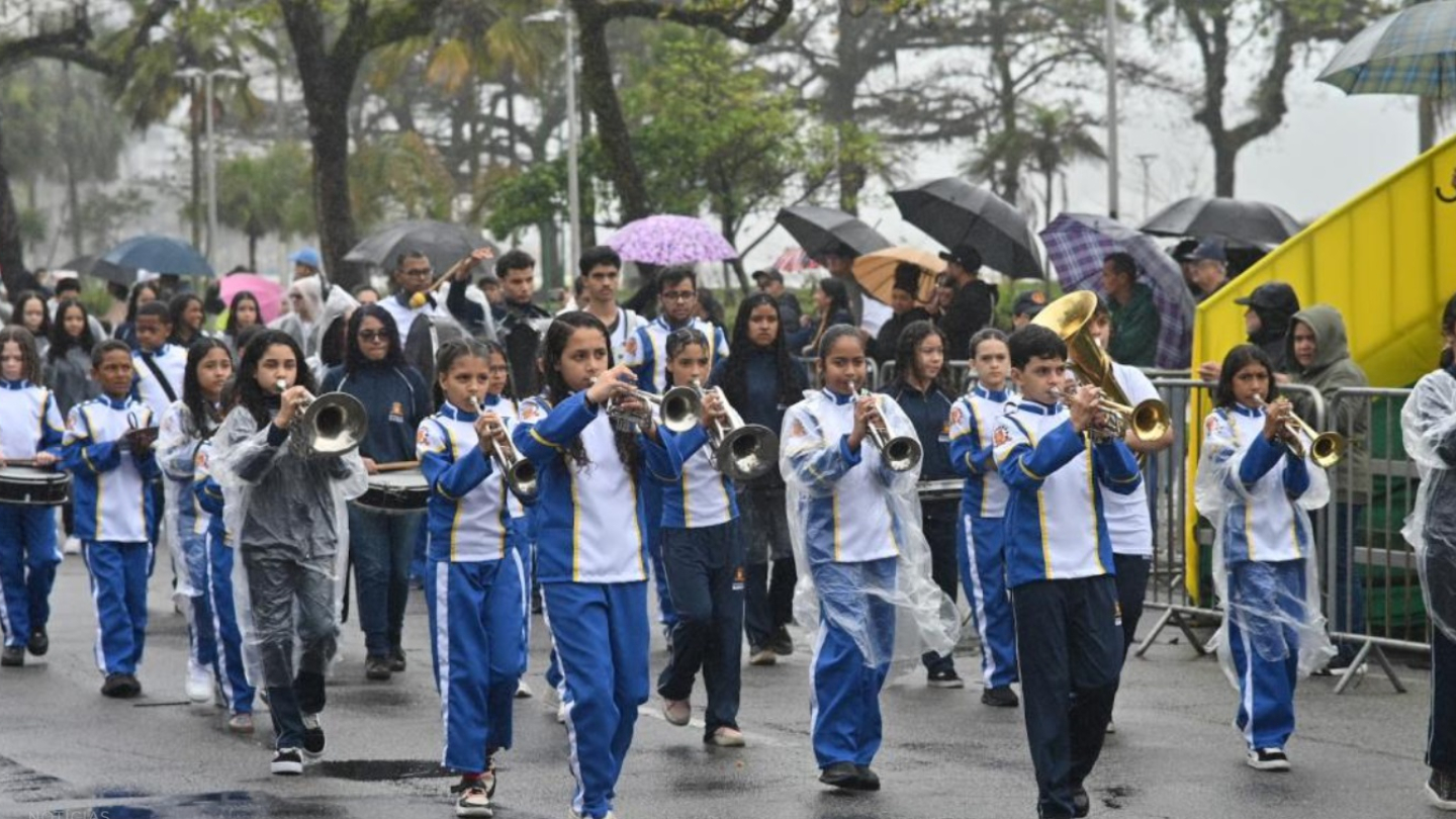 A imagem retrata o desfile de 7 de Setembro em Santos, realizado em um dia chuvoso. Em primeiro plano, jovens músicos marcham tocando instrumentos de sopro e percussão, vestidos com uniformes azul e branco. Alguns usam capas de chuva transparentes para se proteger da garoa. Ao fundo, o público acompanha o evento sob guarda-chuvas coloridos, reforçando o clima úmido do momento.