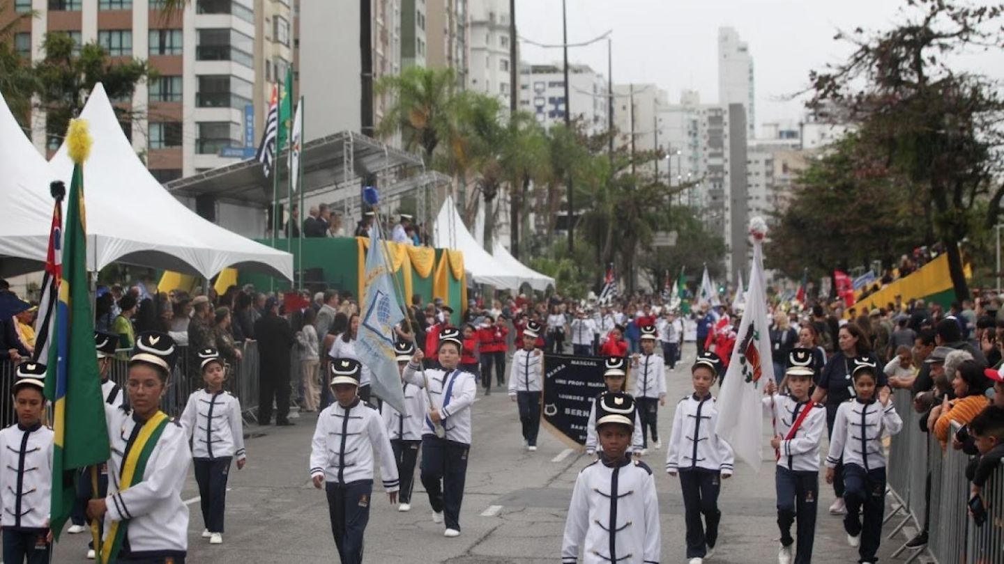 A imagem mostra o desfile cívico de 7 de Setembro em Santos, realizado em uma avenida cercada por prédios e arquibancadas com tendas brancas para autoridades e convidados. O clima está nublado e chuvoso, mas a celebração segue com grande participação popular. Na frente, um grupo de estudantes uniformizados, vestidos de branco com detalhes azuis e chapéus, marcha carregando bandeiras, incluindo a do Brasil, do Estado de São Paulo e outras institucionais.