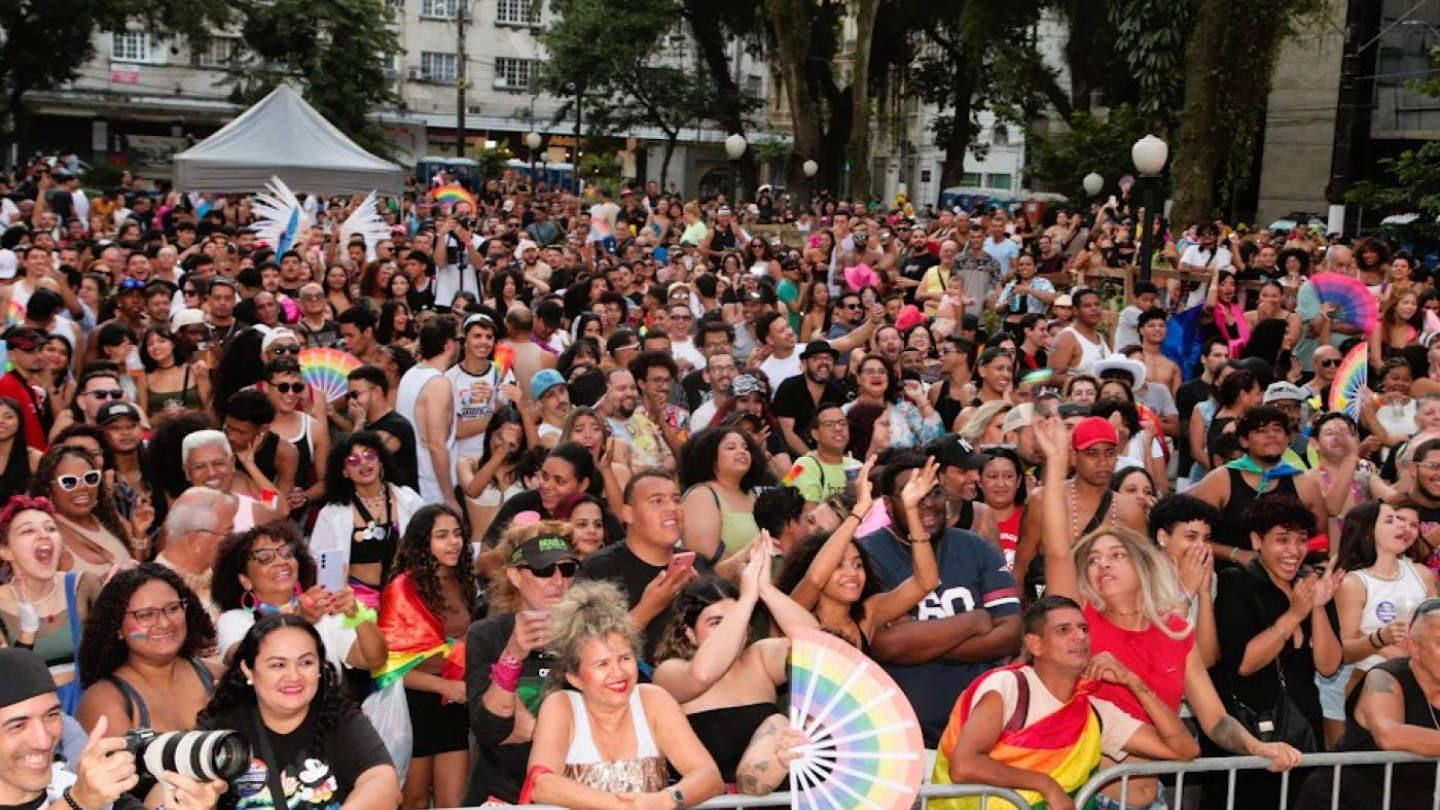 Multidão em evento ao ar livre, com muitas pessoas sorrindo, vibrando e exibindo bandeiras e acessórios coloridos que simbolizam a diversidade