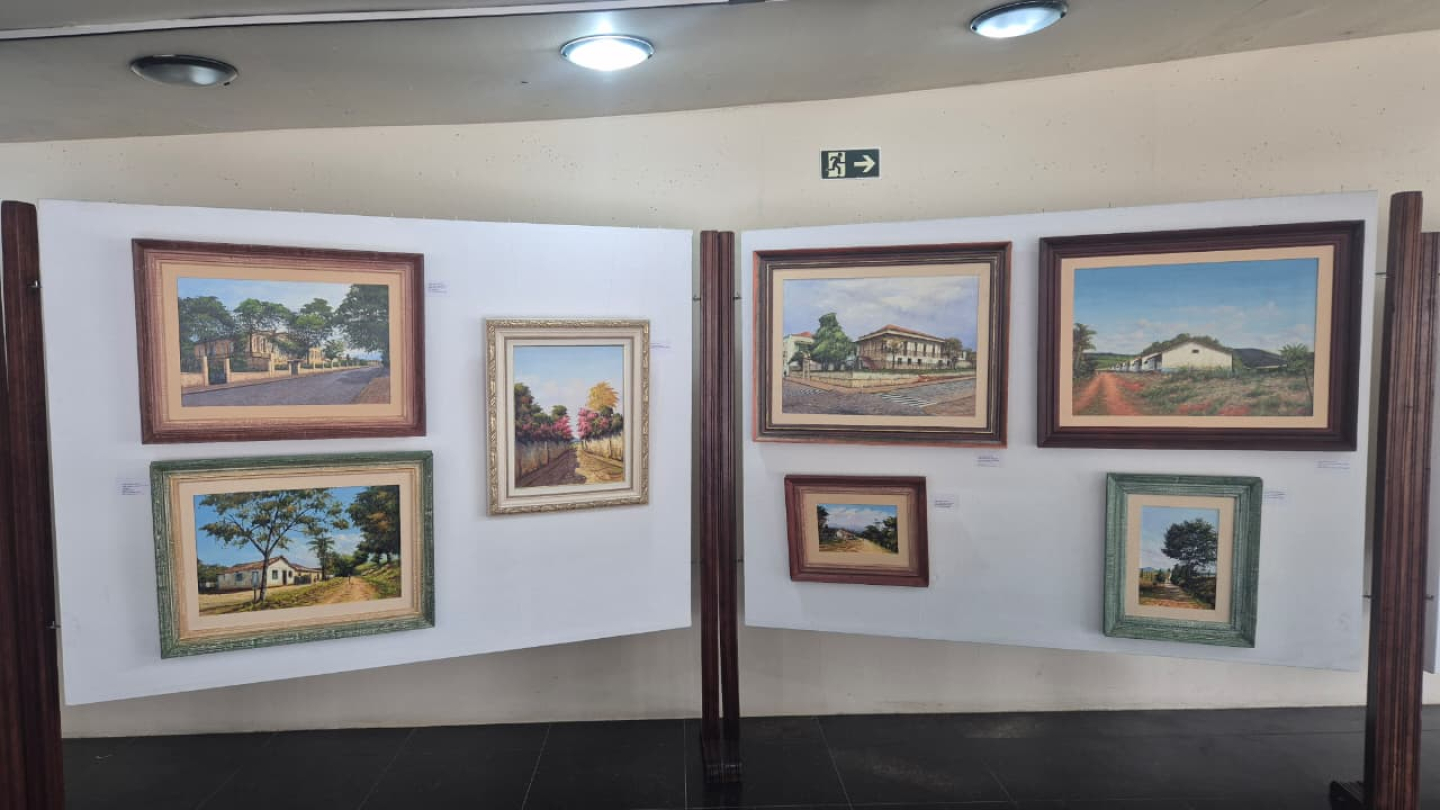 Diversos quadros de pintura, de casas e paisagens, expostos em painéis brancos