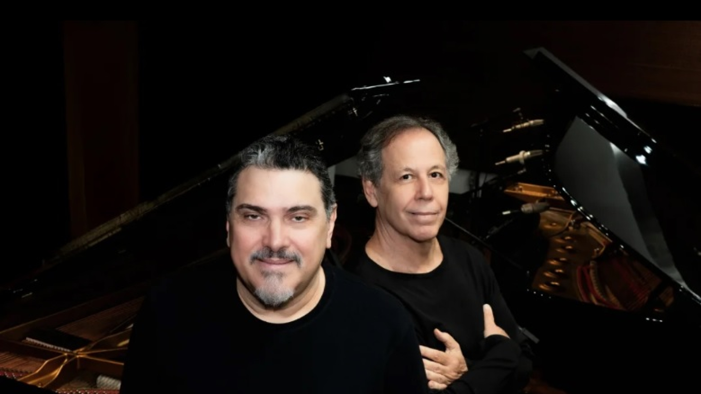 Dois homens, de roupa preta, posicionados lado a lado, em frente a um piano de cauda aberto