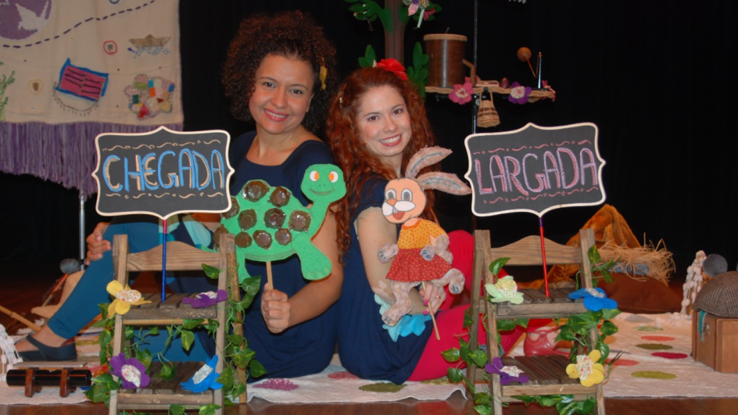 Duas mulheres sentadas lado a lado seguram fantoches de tartaruga e coelho, com placas escritas “CHEGADA” e “LARGADA” em cenário colorido infantil