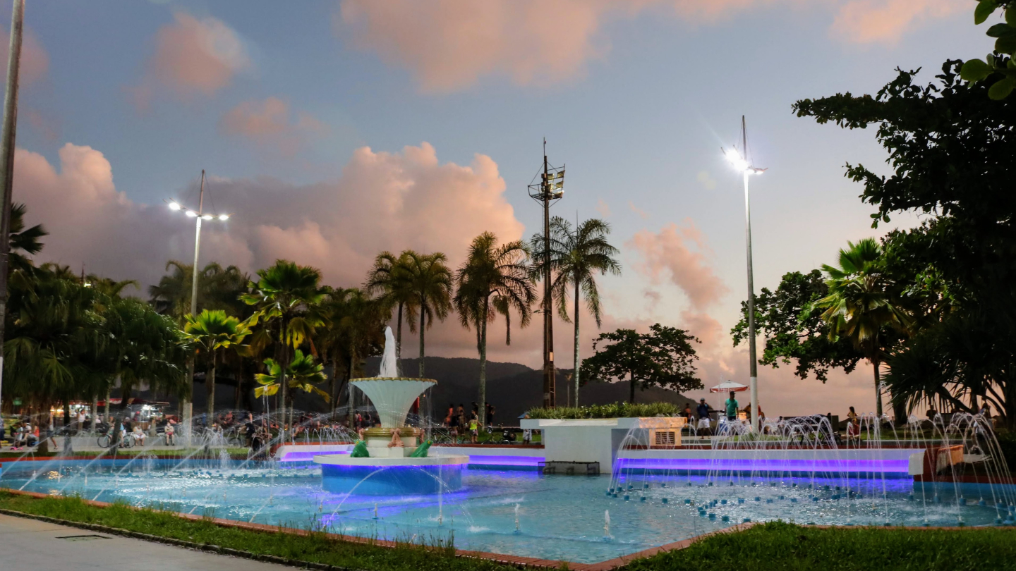 Fonte iluminada com jatos de água, cercada por palmeiras e pessoas ao entardecer