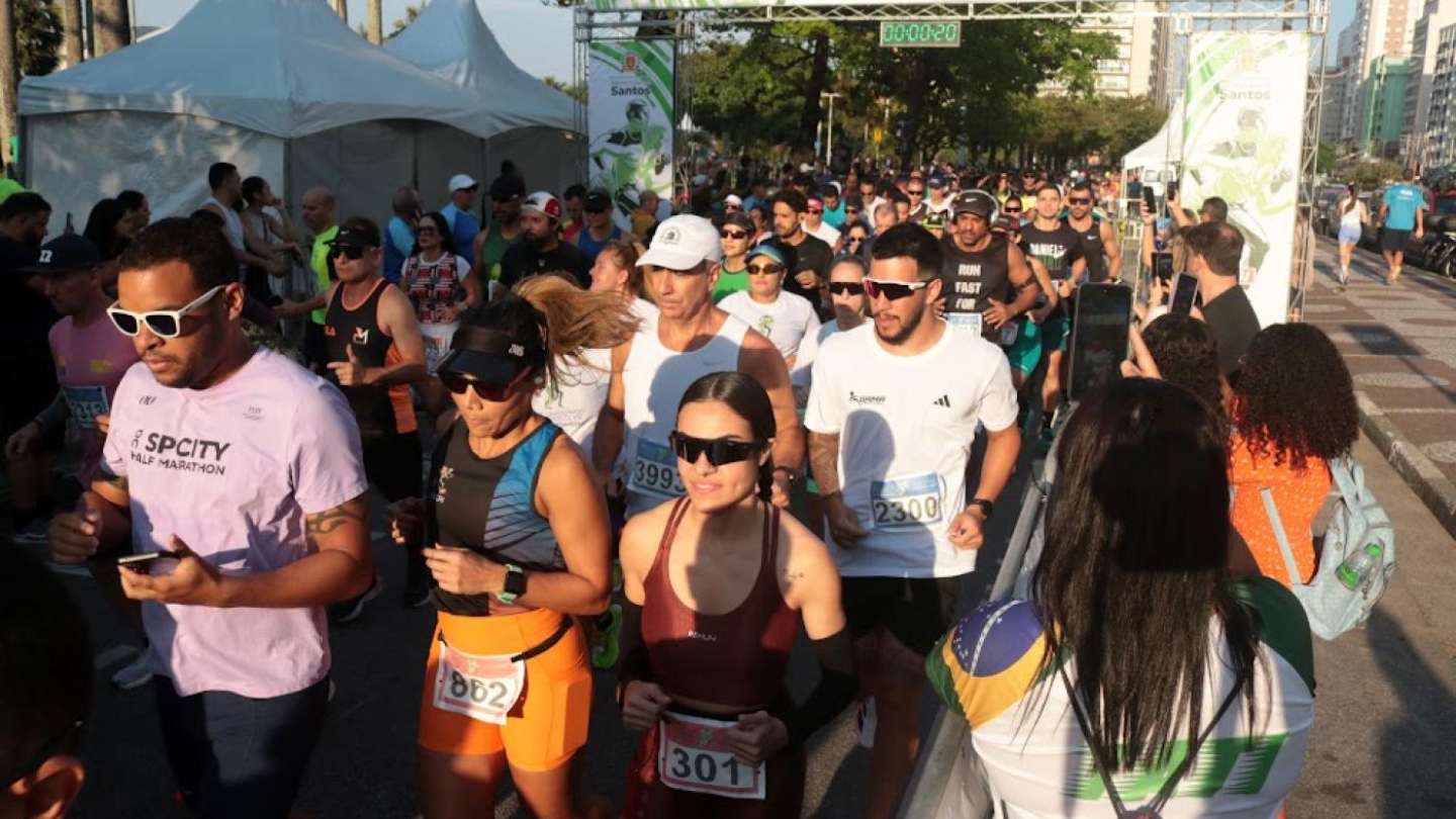 A imagem mostra a largada de uma corrida de rua em Santos, durante o 38º Campeonato Santista de Pedestrianismo. Diversos atletas aparecem concentrados, correndo logo após a partida, com seus números de inscrição fixados nas roupas esportivas. O ambiente é animado, com público acompanhando de perto e registrando o momento com celulares. No fundo, vê-se o pórtico da prova com o cronômetro marcando o início da competição, além de árvores e prédios que compõem a paisagem urbana da cidade.