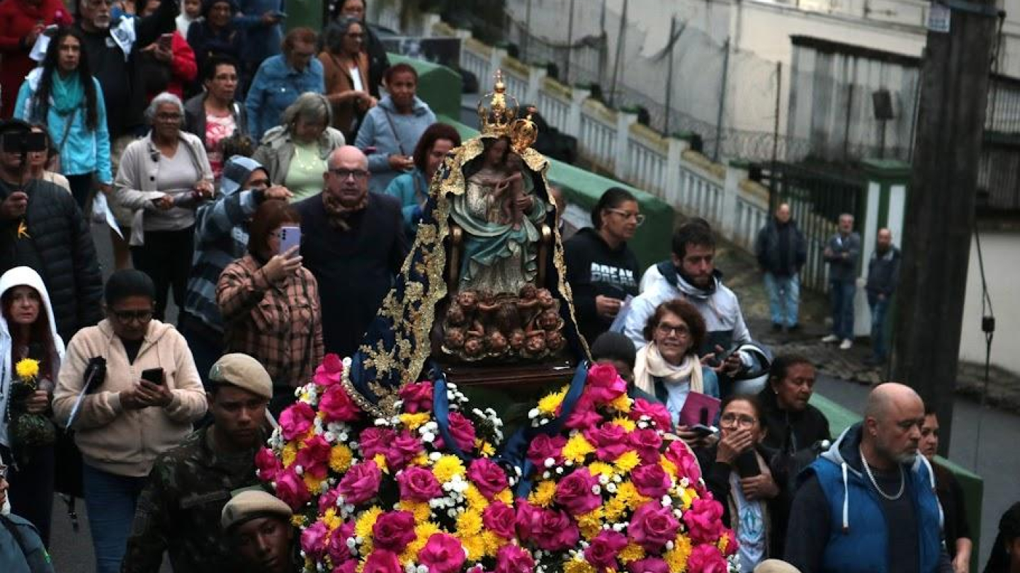 A imagem registra a tradicional procissão de Nossa Senhora do Monte Serrat, padroeira de Santos. Fiéis acompanham a imagem da santa, ricamente ornamentada com um manto bordado e coroada, conduzida em um andor enfeitado com flores coloridas. O cortejo reúne pessoas de diferentes idades, que caminham pelas ruas em demonstração de fé e devoção, muitas delas carregando flores e registrando o momento. A cena traduz a importância cultural e religiosa dessa celebração.