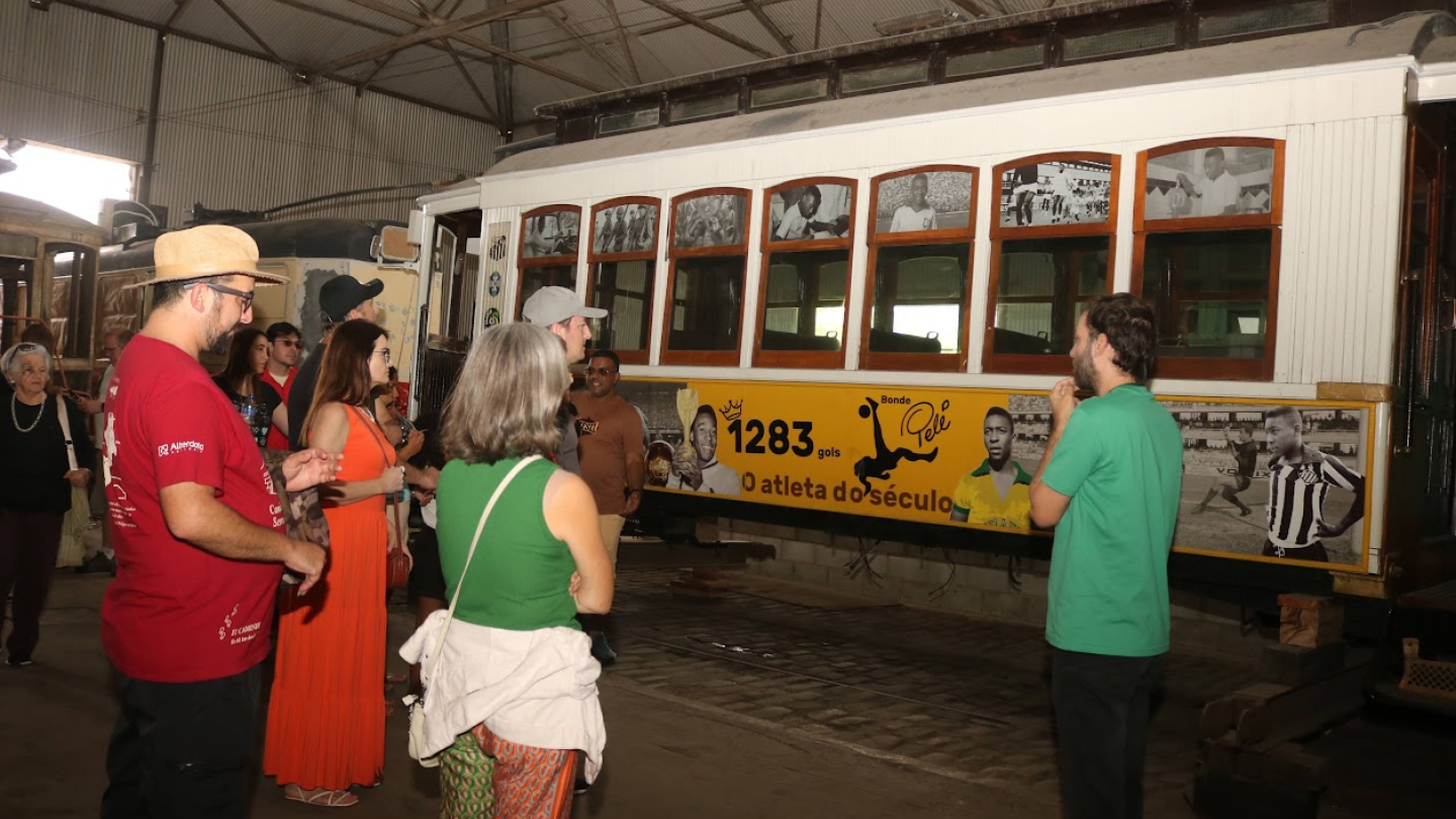 Grupo de pessoas na garagem de bondes, em frente ao Bonde Pelé. Um guia, vestido com uma camisa verde, está de costas, falando com o grupo