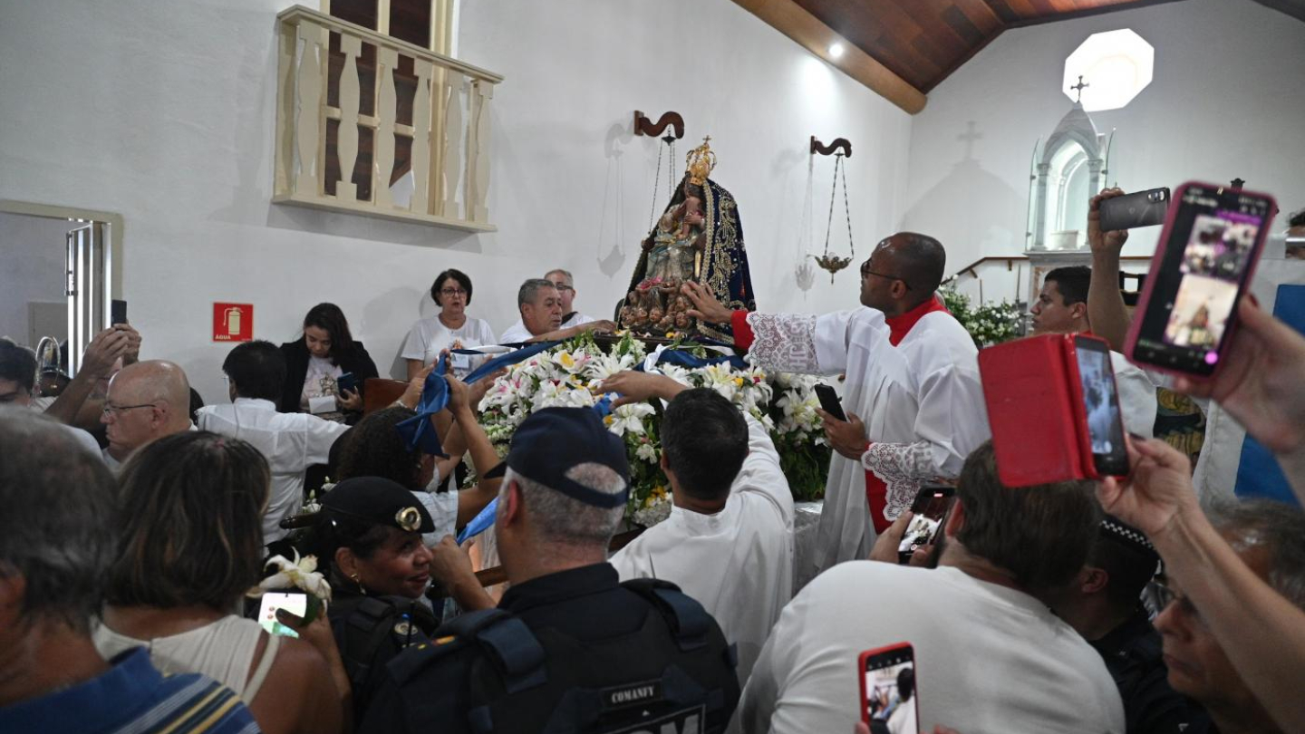 A imagem mostra a celebração do Dia de Nossa Senhora do Monte Serrat, padroeira de Santos. No interior de uma capela, a imagem da santa, ricamente adornada com manto azul bordado e coroa dourada, está sobre um andor ornamentado com flores brancas. Fiéis se aproximam para tocar ou amarrar fitas, demonstrando devoção, enquanto muitos registram o momento com celulares. Ao redor, padres, coroinhas e membros da comunidade participam do ato religioso, que reúne grande número de pessoas em clima de fé e emoção.