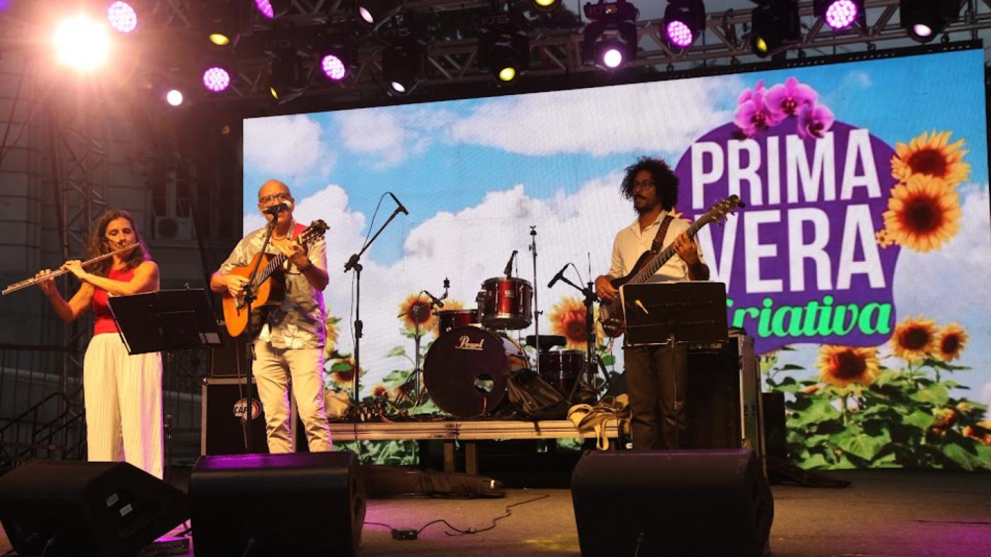 A imagem mostra uma apresentação musical ao vivo durante o evento Primavera Criativa, em Santos. No palco, três músicos tocam seus instrumentos: à esquerda, uma mulher toca flauta transversal; no centro, um homem toca violão e canta; e à direita, outro músico toca baixo elétrico. Ao fundo, um grande telão exibe a identidade visual do evento, decorado com flores como girassóis e orquídeas, reforçando o clima alegre e colorido da primavera. A iluminação do palco, com refletores em tons de roxo e amarelo.