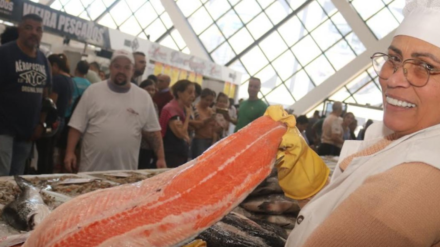 Uma feirante sorridente, usando touca, óculos e luvas amarelas, exibe um grande filé de salmão em uma banca repleta de peixes e frutos do mar. Ao fundo, várias pessoas observam os produtos em um mercado movimentado.