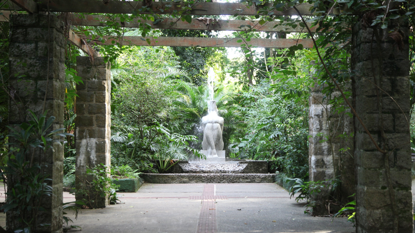 Fonte em forma de escultura feminina cercada por vegetação densa em um jardim tropical