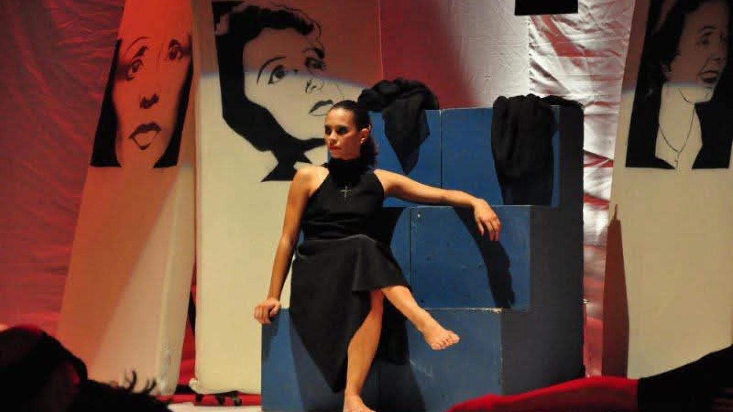 Bailarina de vestido preto sentada em um bloco azul, em cena teatral com painéis ao fundo que exibem rostos desenhados em preto e branco