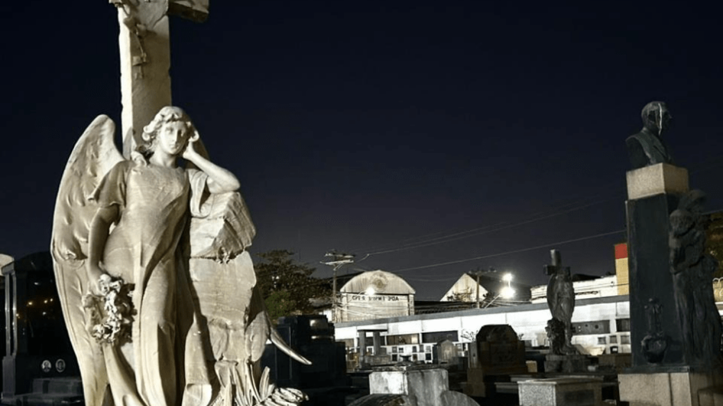 Escultura de anjo, embaixo de uma cruz, ambos feitos em mármore branco, localizada no cemitério do Paquetá