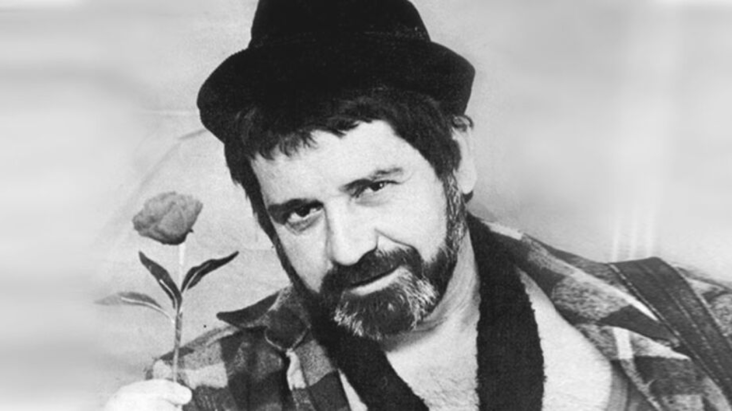 Homem de barba e chapéu, em foto em preto e branco, segura uma flor na mão com expressão serena