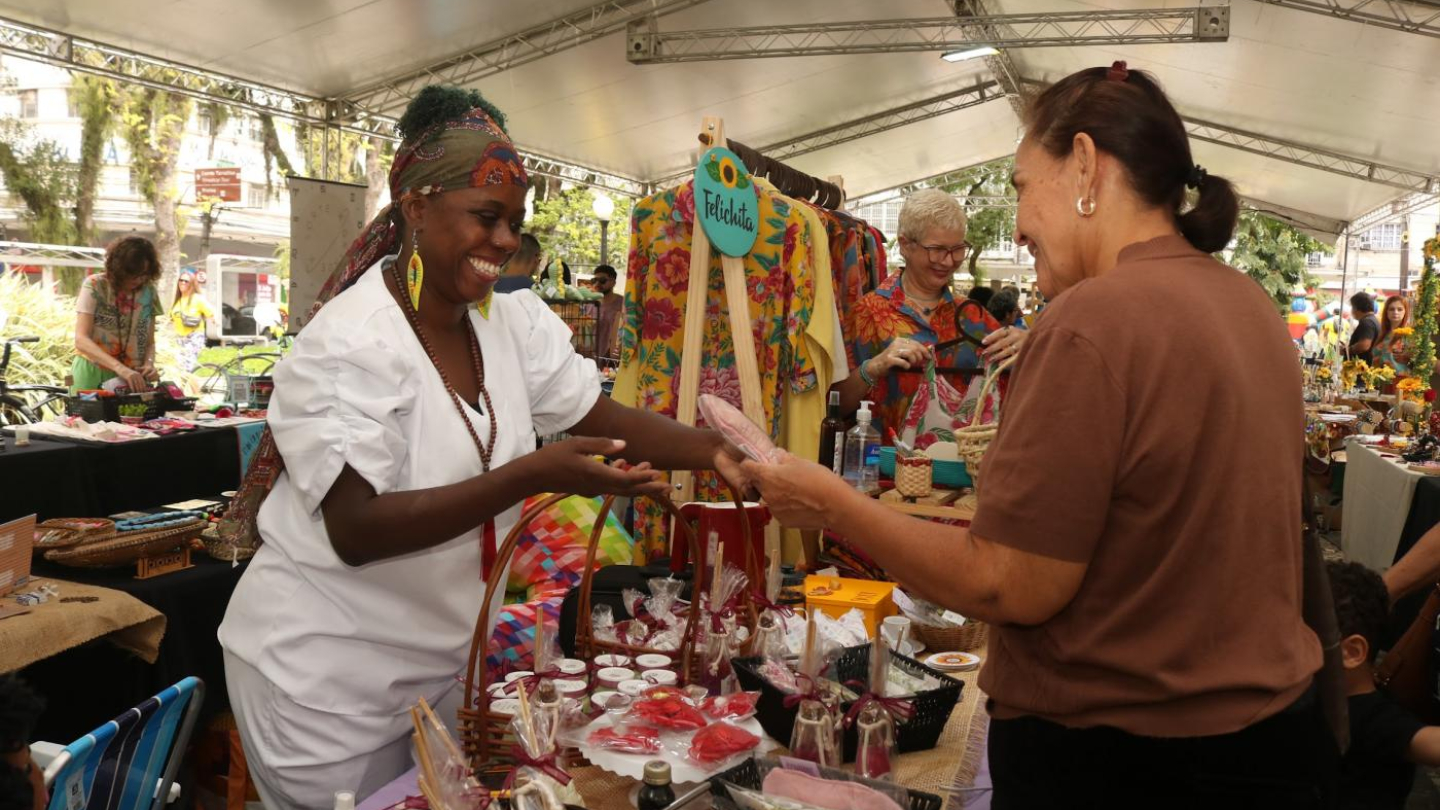 A imagem mostra uma cena alegre em uma feira cultural realizada ao ar livre, sob uma grande tenda. Em primeiro plano, uma expositora sorridente, vestida de branco e com adereços coloridos, atende uma visitante que observa e compra seus produtos. Sobre a mesa, há diversos itens artesanais, como sabonetes, óleos, enfeites e lembranças embaladas com capricho.