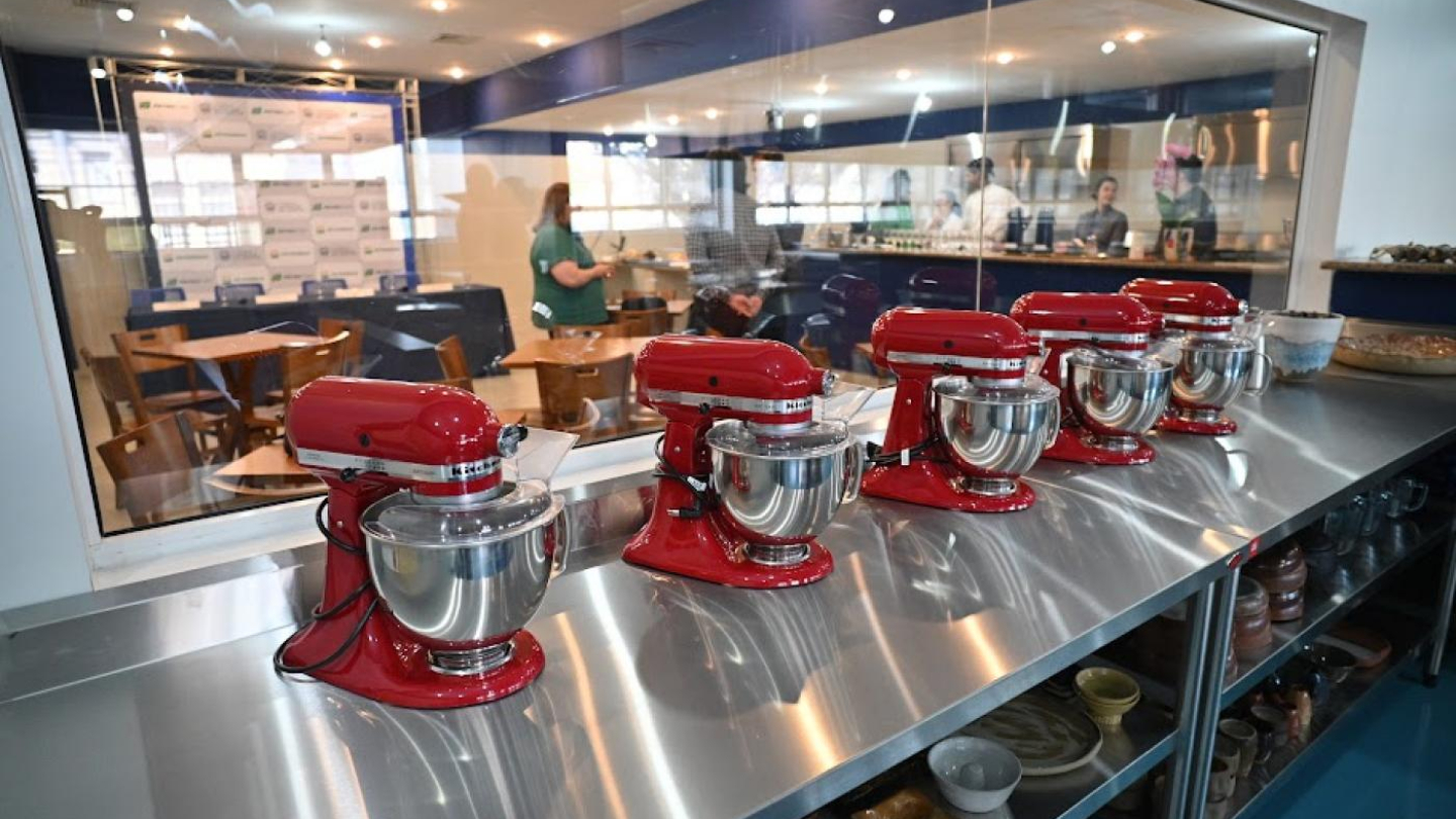 Na imagem vemos uma bancada de aço inoxidável onde estão dispostas cinco batedeiras planetárias vermelhas, todas alinhadas em fila. Através do vidro ao fundo, é possível observar uma cozinha-escola ou laboratório de gastronomia, com algumas pessoas em atividade, incluindo chefs e participantes. O ambiente é moderno, bem iluminado e organizado, transmitindo a ideia de um espaço voltado para aulas práticas ou demonstrações culinárias.