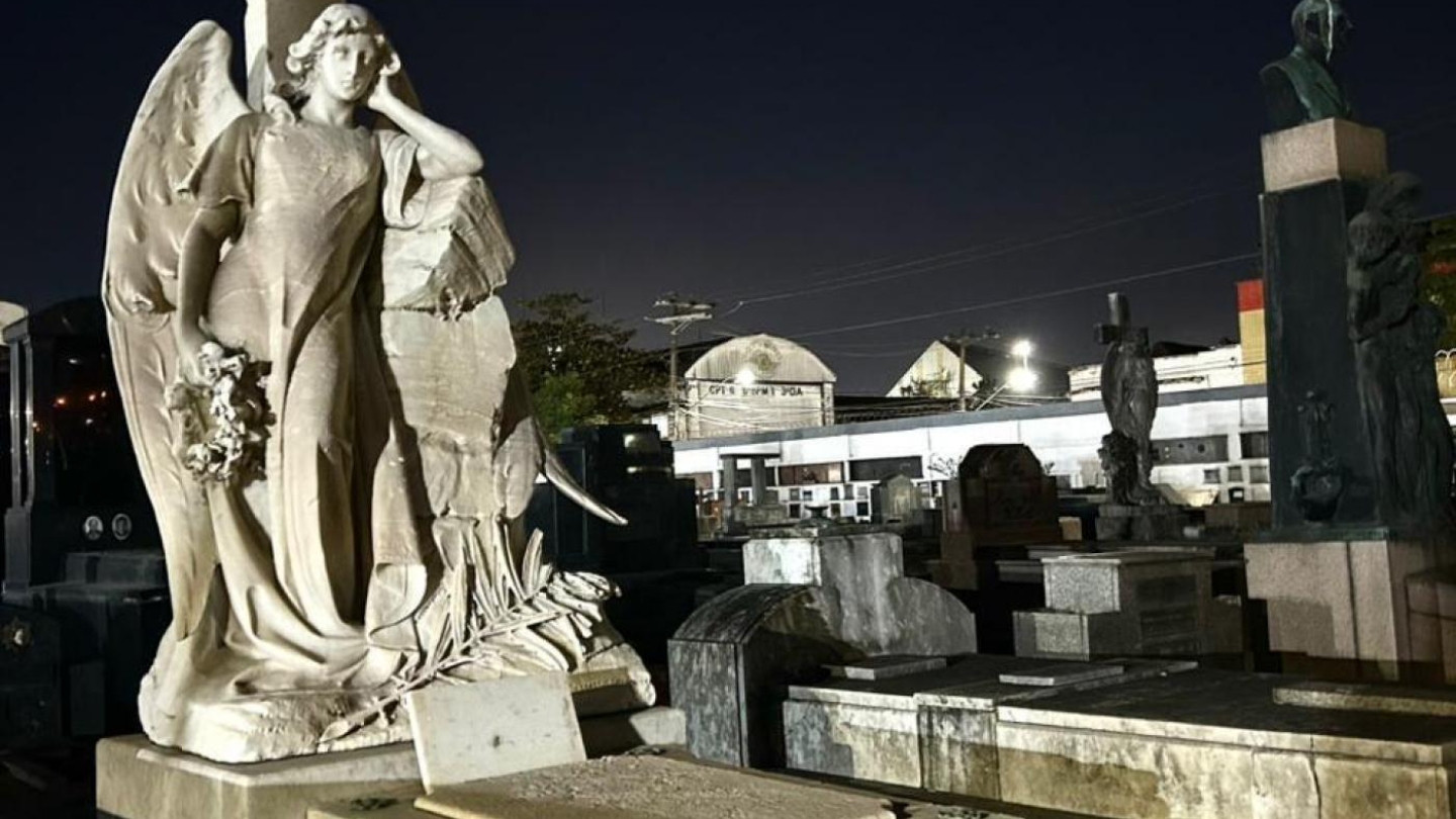 A imagem mostra uma cena noturna no Cemitério do Paquetá, em Santos, registrada durante um passeio guiado. Em primeiro plano, destaca-se uma imponente escultura de anjo em mármore claro, em posição contemplativa ao lado de uma grande cruz ornamentada. O anjo segura flores, símbolo de luto e memória, enquanto sua expressão serena transmite paz. O túmulo onde está a escultura é robusto, em pedra clara, com inscrições na lápide.