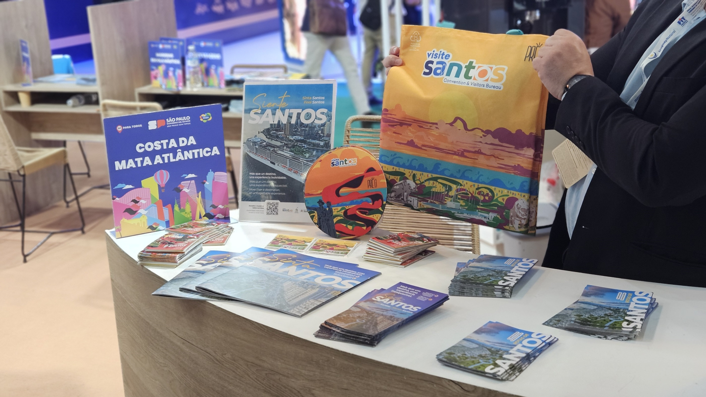 A imagem mostra um estande de divulgação turística com materiais promocionais da cidade de Santos, em um ambiente de feira ou evento. Sobre um balcão de madeira clara estão organizados diversos folhetos, revistas e folders coloridos com informações turísticas.