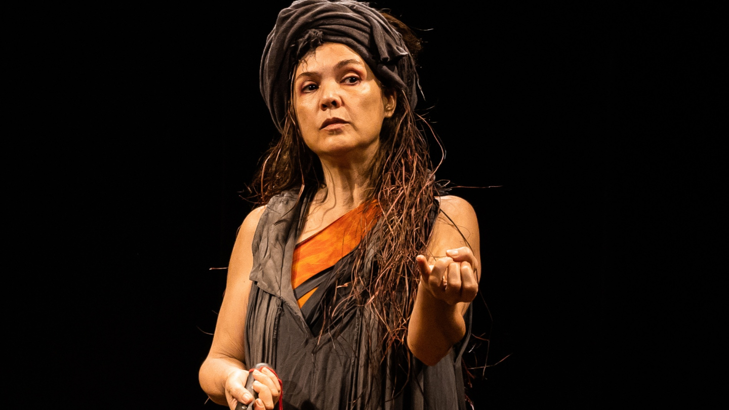 Mulher em traje cerimonial ou teatral, com turbante de tecido e roupa escura com detalhe laranja, gesticula sob fundo preto em cena de forte expressão dramática