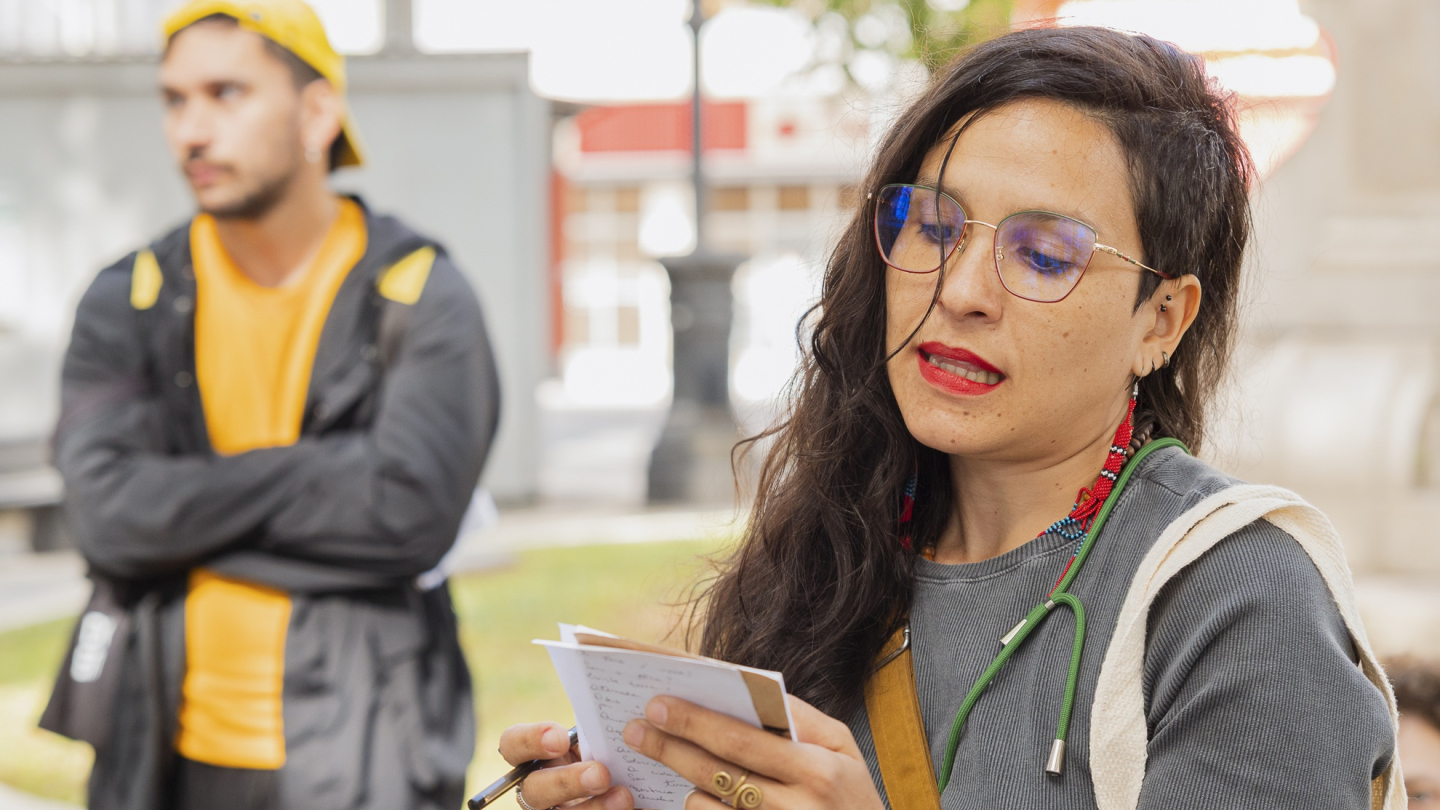 Mulher de óculos e batom vermelho segura um caderno e caneta, aparentando fazer anotações, enquanto outra pessoa está desfocada ao fundo