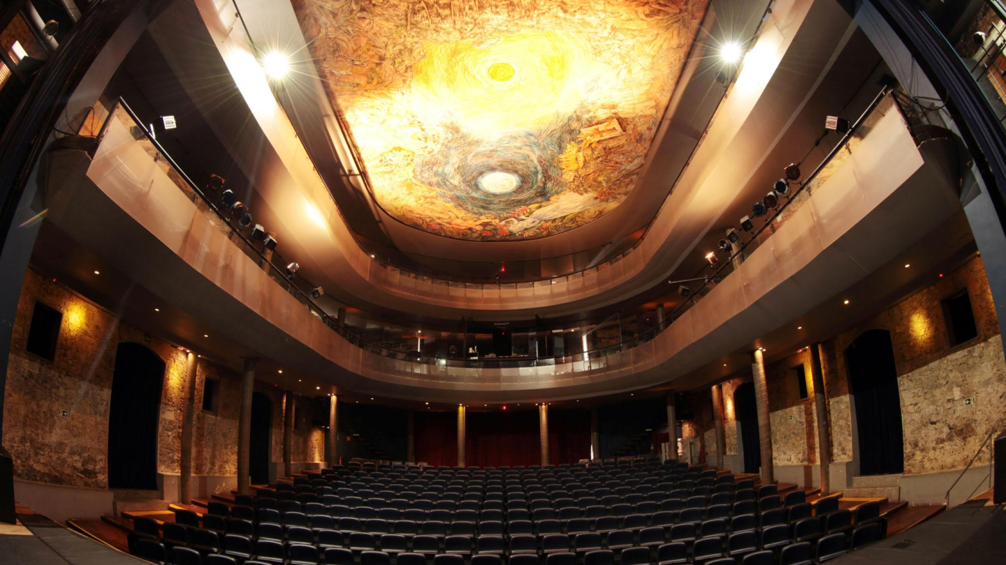 Interior de teatro com plateia vazia, dois andares de balcões e teto decorado com uma grande pintura iluminada