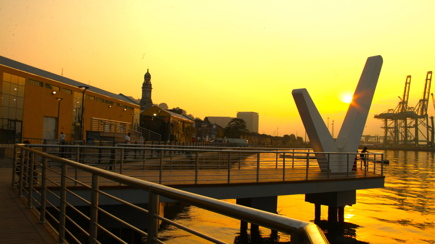 A imagem mostra o pôr do sol no Parque Valongo, em Santos, com o céu em tons dourados refletindo sobre as águas do estuário. Em primeiro plano, aparece a estrutura metálica do deque, com guarda-corpos de aço e piso de madeira, onde algumas pessoas observam a paisagem. À esquerda, o prédio amarelo do complexo cultural e turístico se destaca, e ao fundo é possível ver a torre da Igreja do Valongo e, à direita, os guindastes do porto.