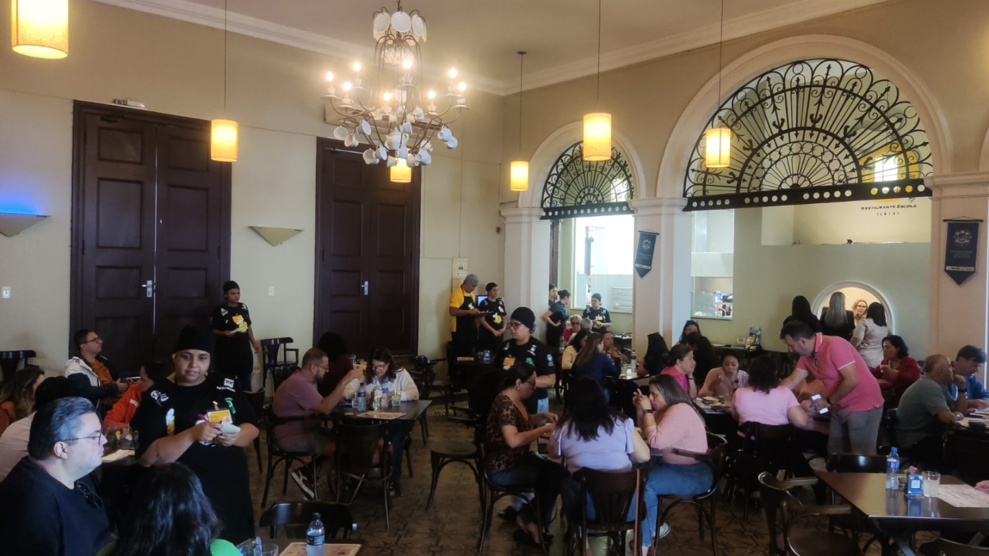 A imagem mostra um ambiente interno movimentado de um restaurante, com várias mesas ocupadas por clientes. O espaço é amplo e elegante, com pé-direito alto, lustres clássicos e luminárias pendentes que criam uma iluminação aconchegante. O piso tem ladrilhos decorativos e as paredes claras contrastam com as grandes portas de madeira escura. Ao fundo, há um balcão e uma cozinha visível através de arcos adornados por grades metálicas ornamentadas.