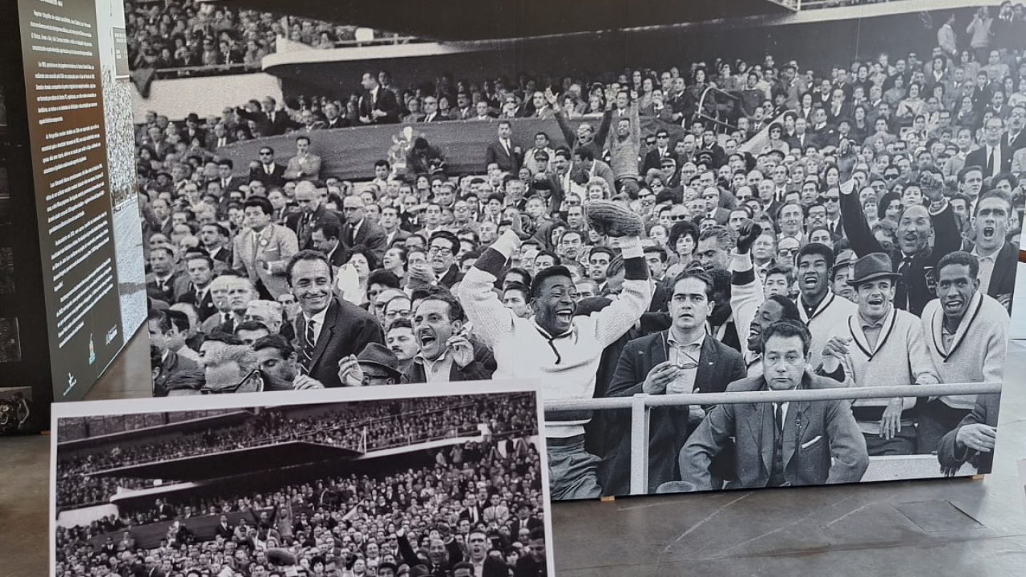 A imagem mostra uma exposição em ambiente interno, com painéis e fotografias em preto e branco. O destaque é uma grande foto histórica que retrata uma multidão em um estádio de futebol, celebrando animadamente. No centro, aparece um jogador sorridente, de agasalho claro, erguendo uma bola, rodeado por companheiros de equipe e torcedores, transmitindo um clima de euforia e conquista.