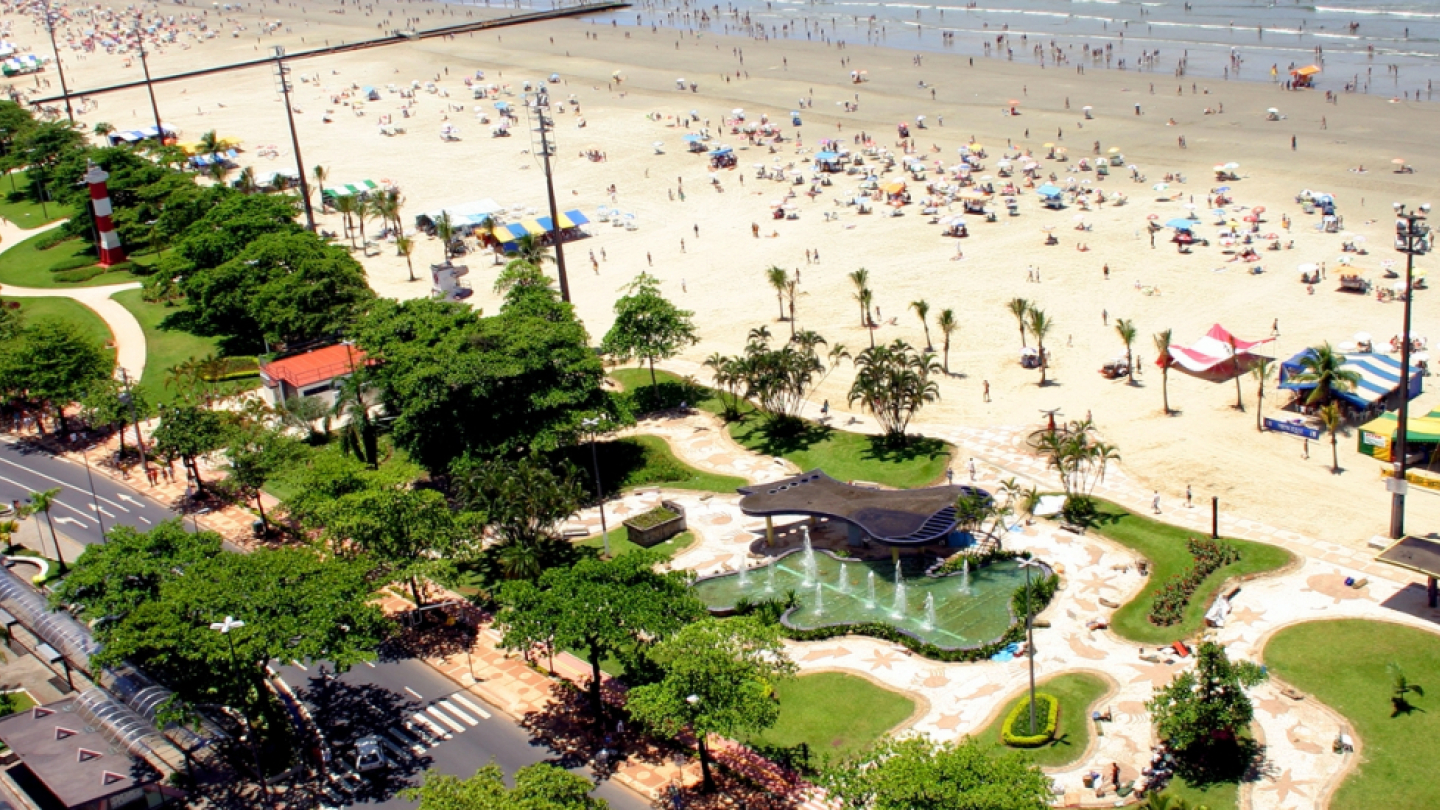 A imagem mostra um trecho da orla de Santos, no litoral paulista, em um dia ensolarado e movimentado. À direita, a praia está repleta de banhistas, guarda-sóis coloridos e barracas, com o mar ao fundo e ondas suaves. No centro, destaca-se o jardim da orla, com caminhos sinuosos, áreas gramadas, palmeiras e uma fonte ornamental com esculturas modernas. À esquerda, vê-se a movimentada avenida da praia, com fluxo de carros, ônibus e ciclovia. Um pequeno farol vermelho e branco é visível entre as árvores.