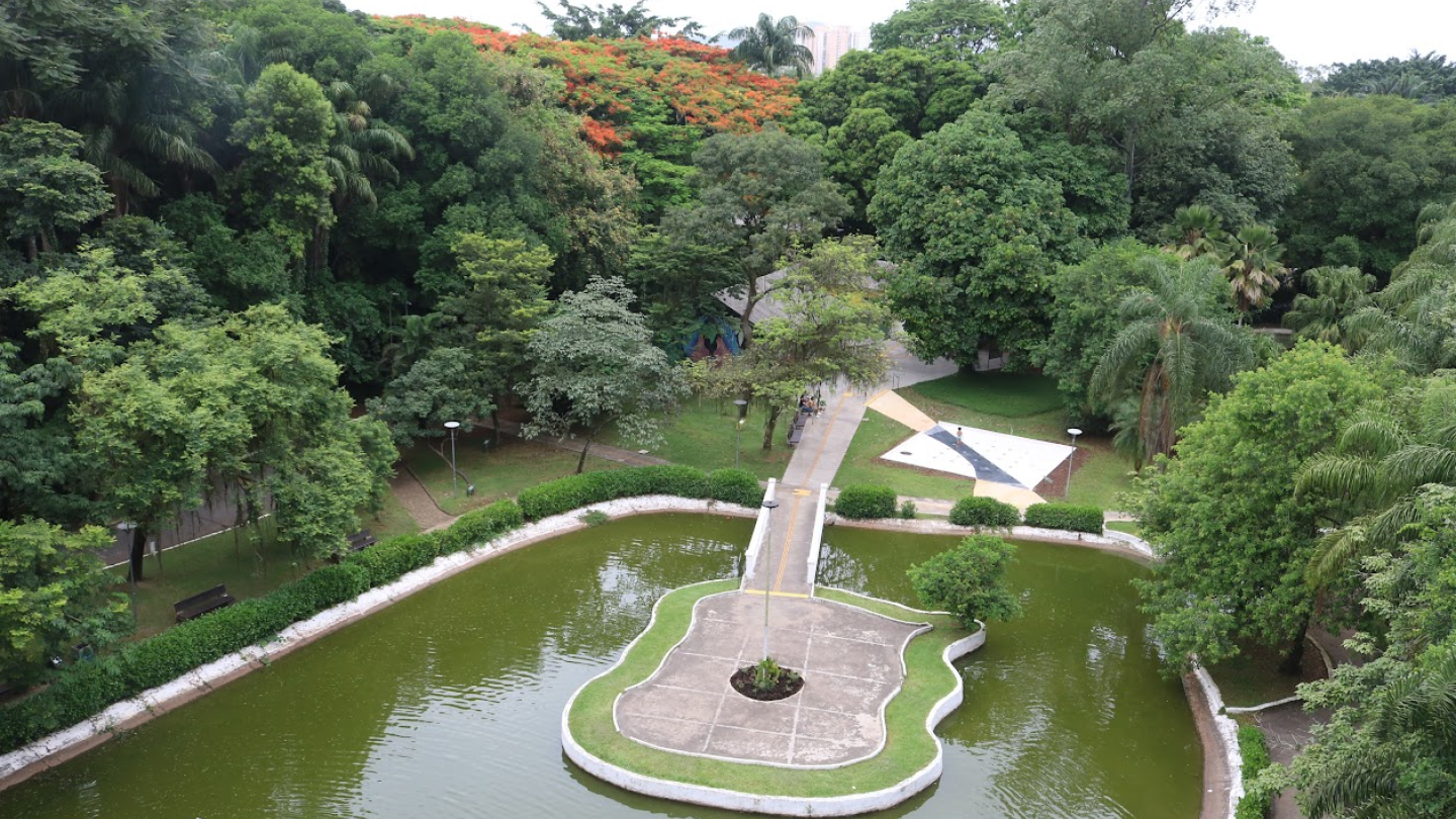 Vista aérea de um parque arborizado com lago em formato de violão. Uma passarela conecta o "corpo do violão" ao restante do parque. Ao fundo, há muitas árvores de diferentes tons de verde e algumas com flores alaranjadas. Caminhos pavimentados cruzam o parque, que também possui áreas gramadas e bancos. Ao centro da imagem, destaca-se o formato do violão desenhado no concreto da ilha.