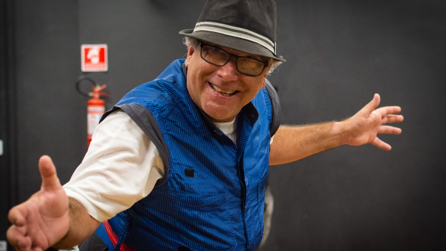Homem sorridente com chapéu e colete azul faz pose animada com os braços abertos em ambiente interno