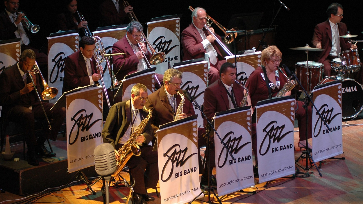 Uma fotografia de uma Big Band, provavelmente uma orquestra de jazz, tocando em um palco. Os músicos estão vestidos de terno e em um fundo escuro