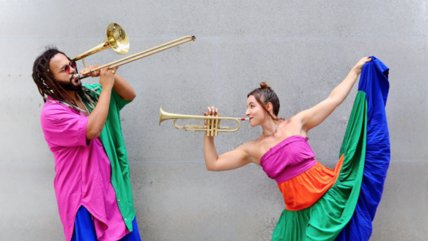 Um homem tocando trombone e uma mullher tocando trompete, ambos de roupas coloridas