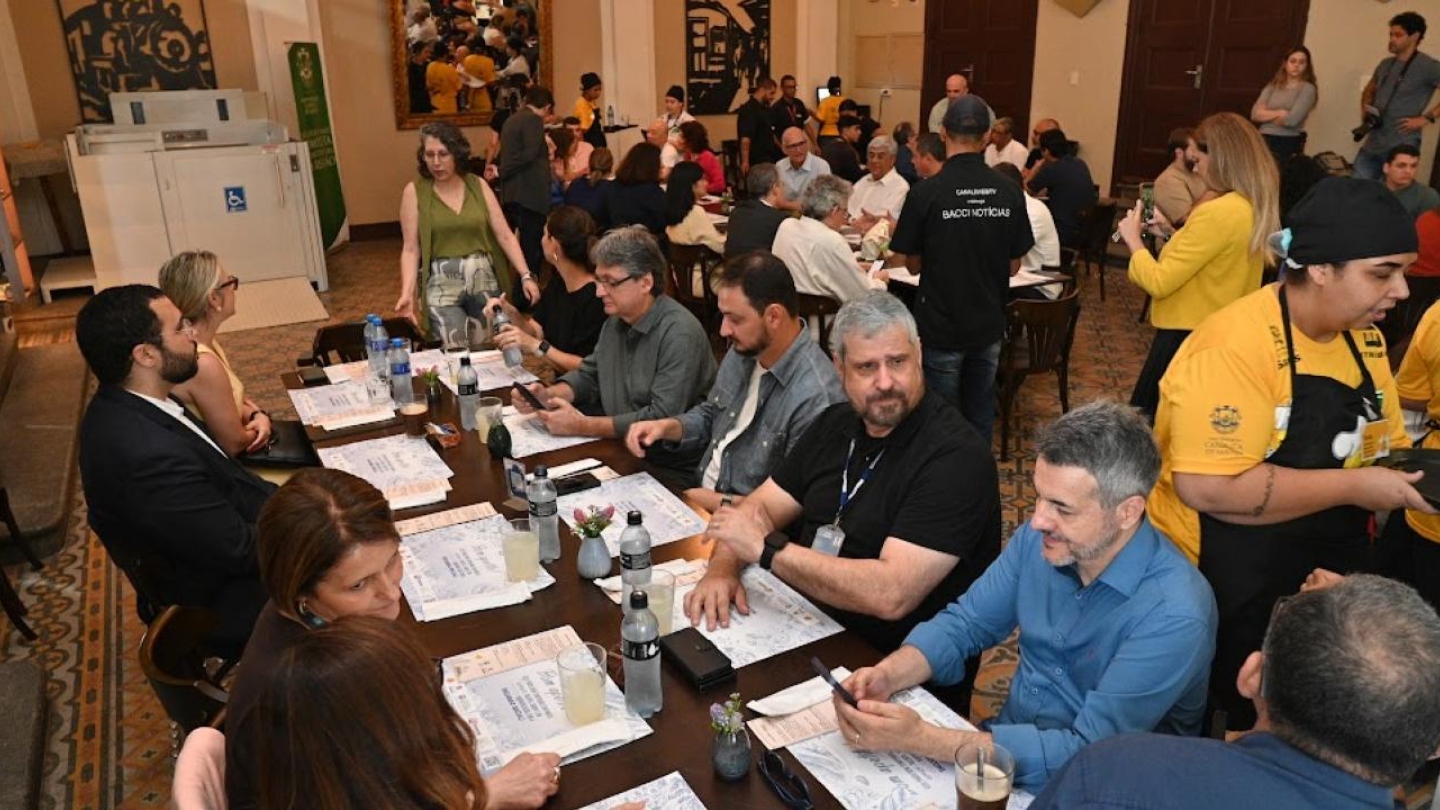 A foto mostra um ambiente movimentado e acolhedor de um restaurante, onde diversas pessoas participam de um almoço coletivo. As mesas estão organizadas lado a lado, com toalhas claras, cardápios e pequenas flores como decoração. Em primeiro plano, um grupo de participantes conversa e manuseia celulares enquanto aguarda o serviço, transmitindo um clima descontraído e de confraternização. Ao fundo, garçons e estudantes uniformizados com camisetas amarelas e aventais pretos circulam entre as mesas.