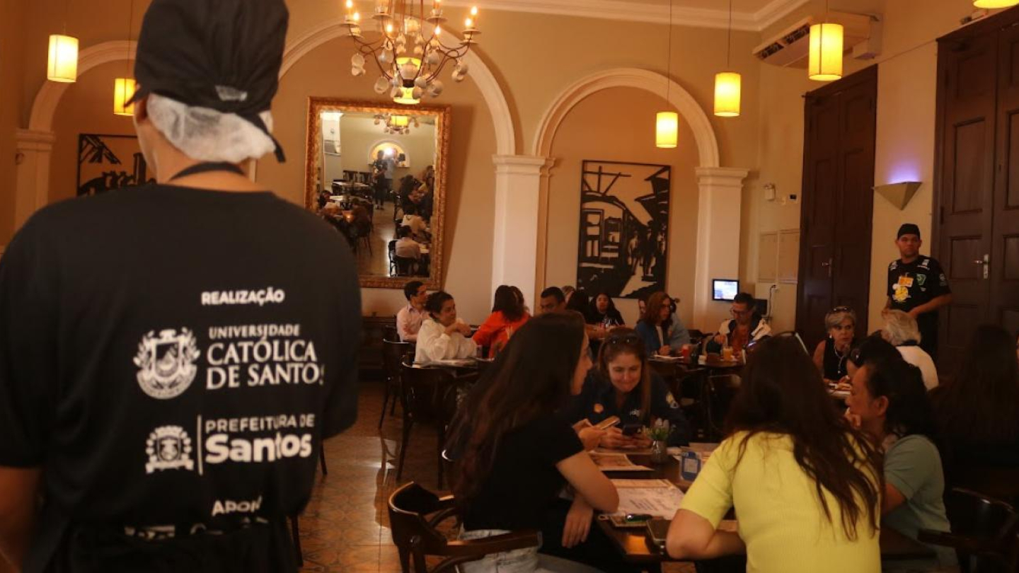 Um ambiente interno de restaurante movimentado, com várias mesas ocupadas por clientes que conversam e fazem suas refeições. O local tem uma atmosfera aconchegante e elegante, com lustres pendentes, espelhos amplos e portas de madeira alta. Em primeiro plano, há uma pessoa de uniforme preto e touca de cozinha, de costas, com a inscrição “Universidade Católica de Santos” e “Prefeitura de Santos” na camiseta, indicando uma parceria institucional.