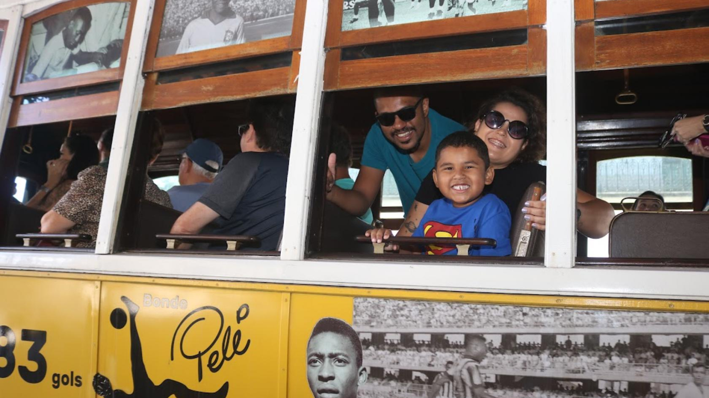 A imagem mostra um bonde temático dedicado a Pelé, com fotografias históricas do Rei do Futebol estampadas na parte externa e também próximas às janelas internas do veículo. Na lateral, aparece uma grande foto de Pelé em destaque, além de ilustrações relacionadas a seus feitos esportivos.